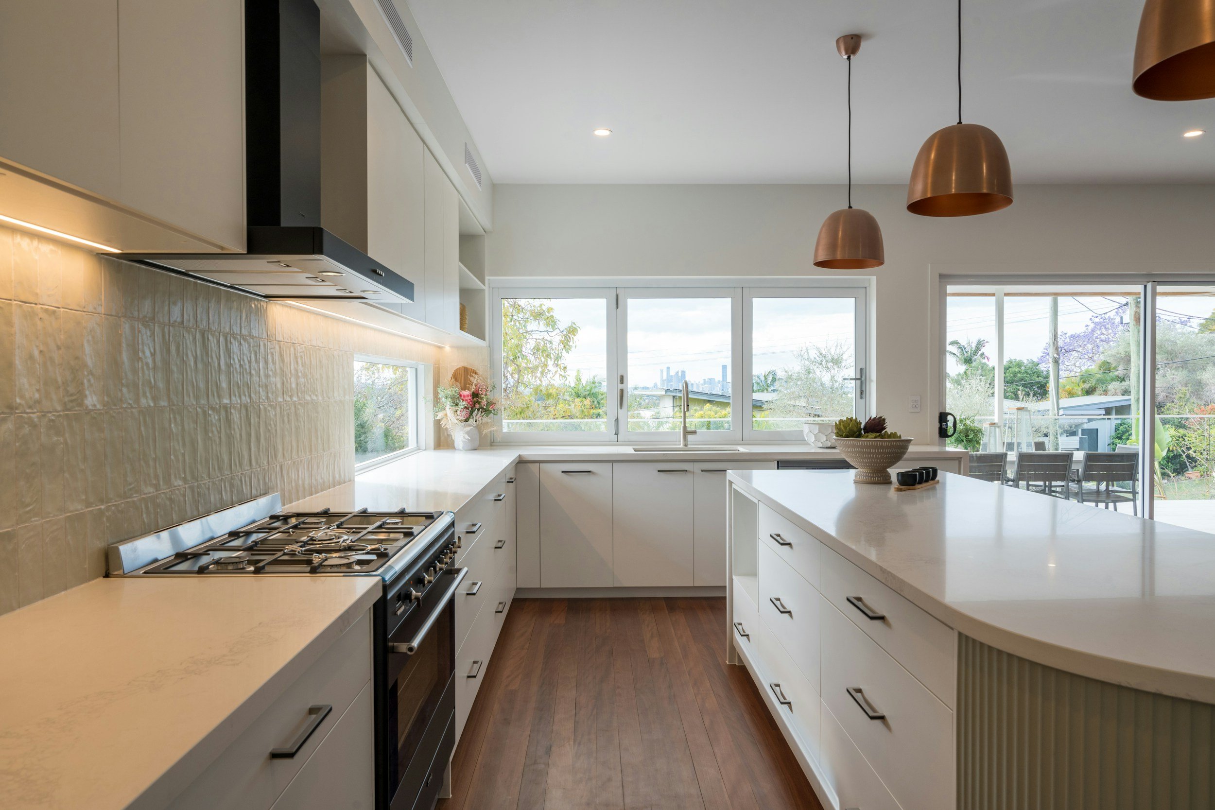 Modern white kitchen with large windows, wooden floors, pendant lights, and outdoor patio view.