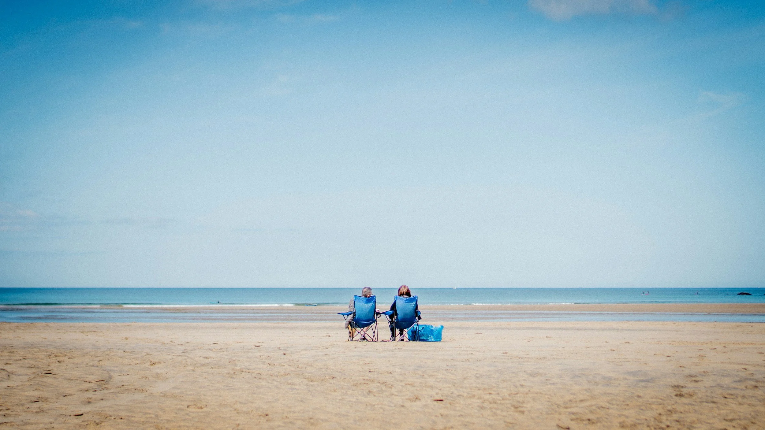 Two people sitting in blue beach chairs on the sandy beach, facing the ocean under a partly cloudy blue sky.