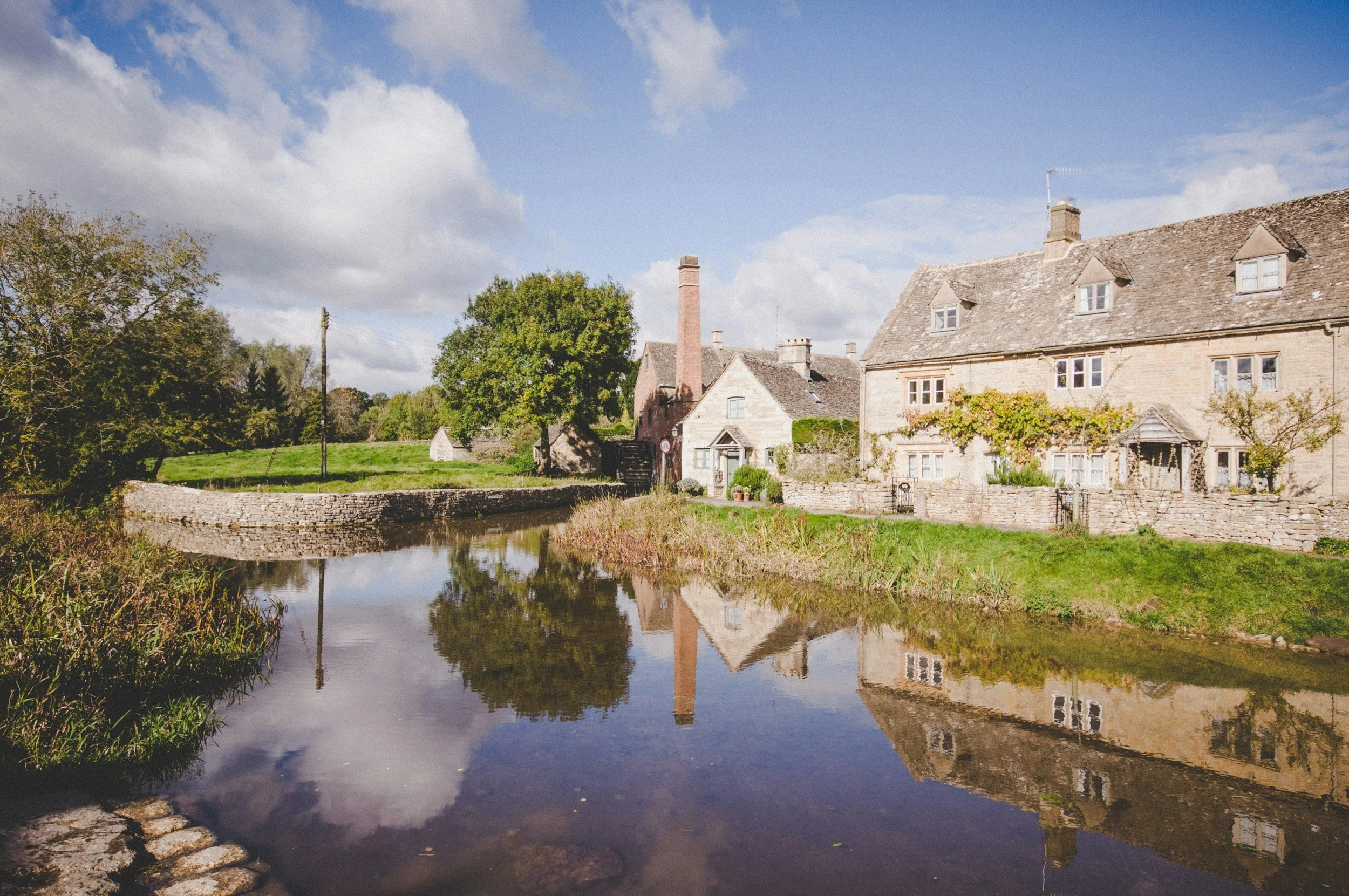 Scenic view of a small river with stone lining, reflecting houses and trees on a sunny day with some clouds.
