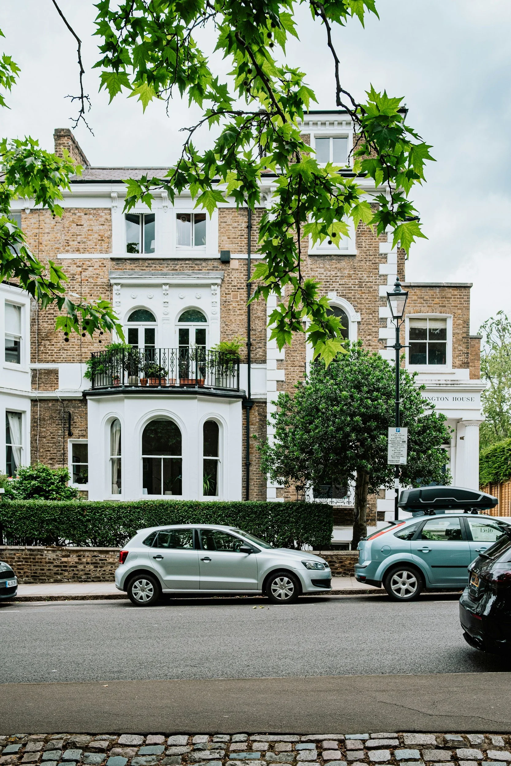 A row of parked cars along a street in front of a large brick building with white decorative accents and multiple windows, partially obscured by green trees with large leaves.