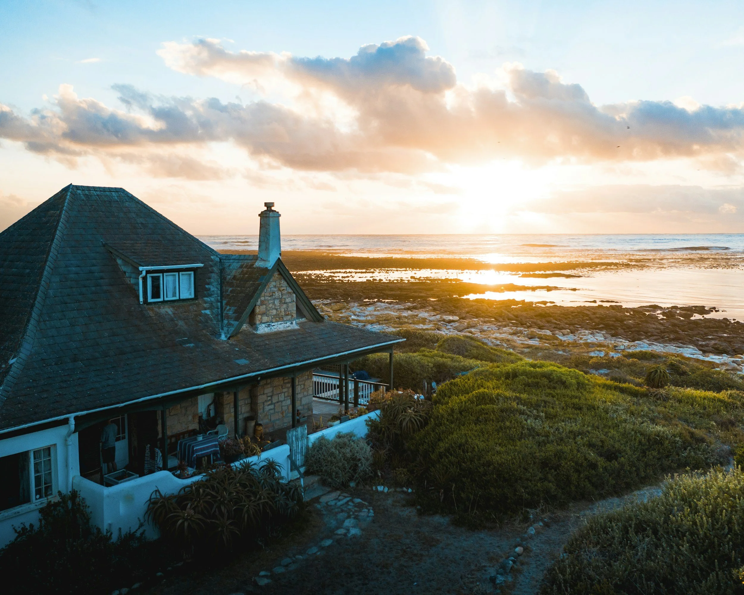 A house with a steep tile roof and a chimney, overlooking a beach with the ocean and a sunset sky with clouds.