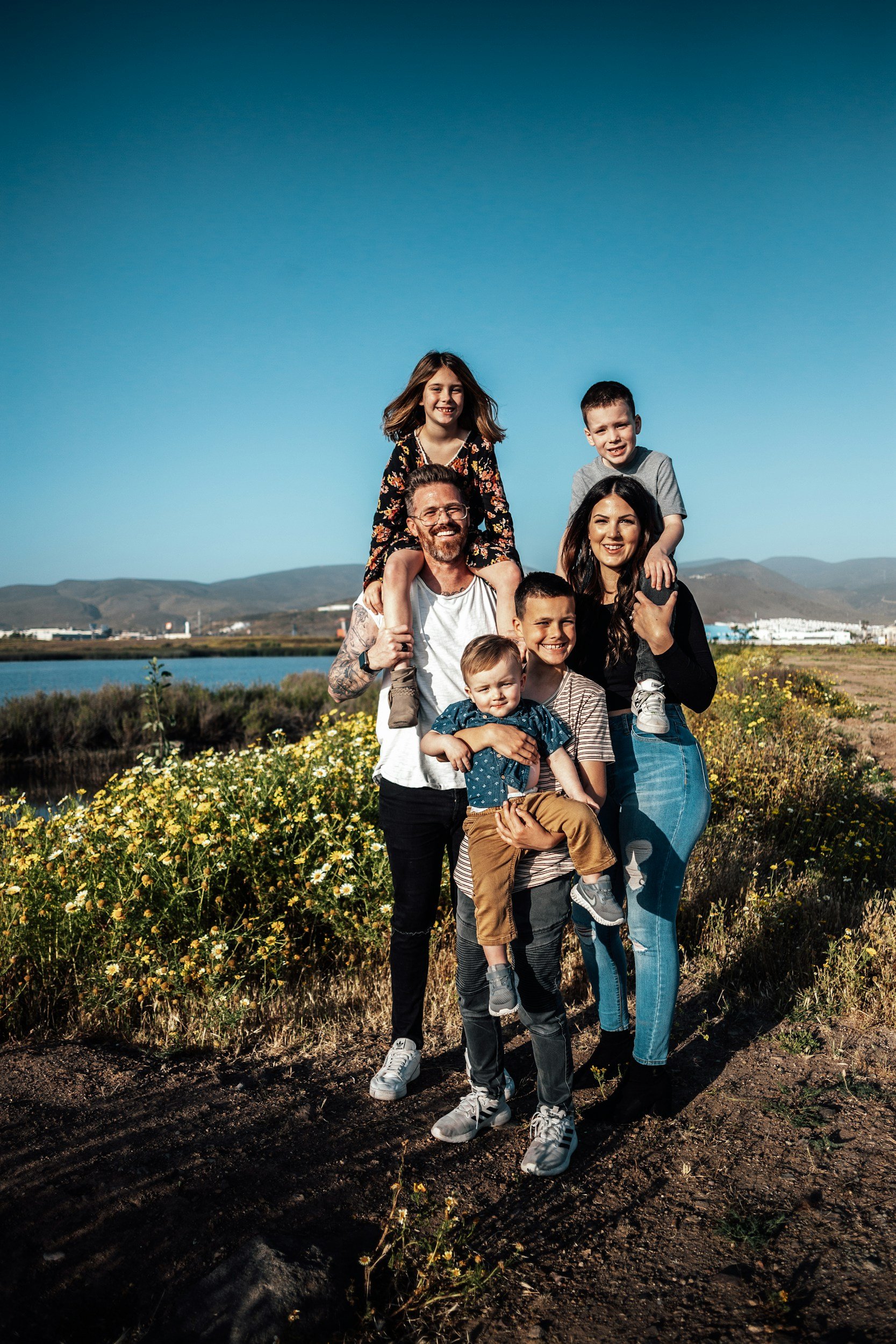 A family of six people outdoors near a body of water, with mountains in the background. They are smiling and posing for the photo, with two children on adults' shoulders, in bright and casual clothing.