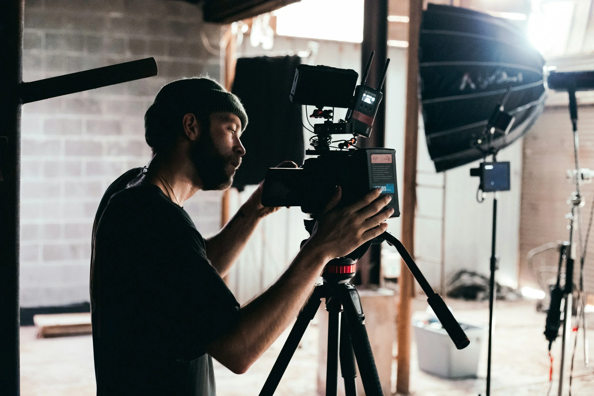 A man wearing a beanie operating a professional camera on a tripod, with studio lighting equipment and a boat in the background.