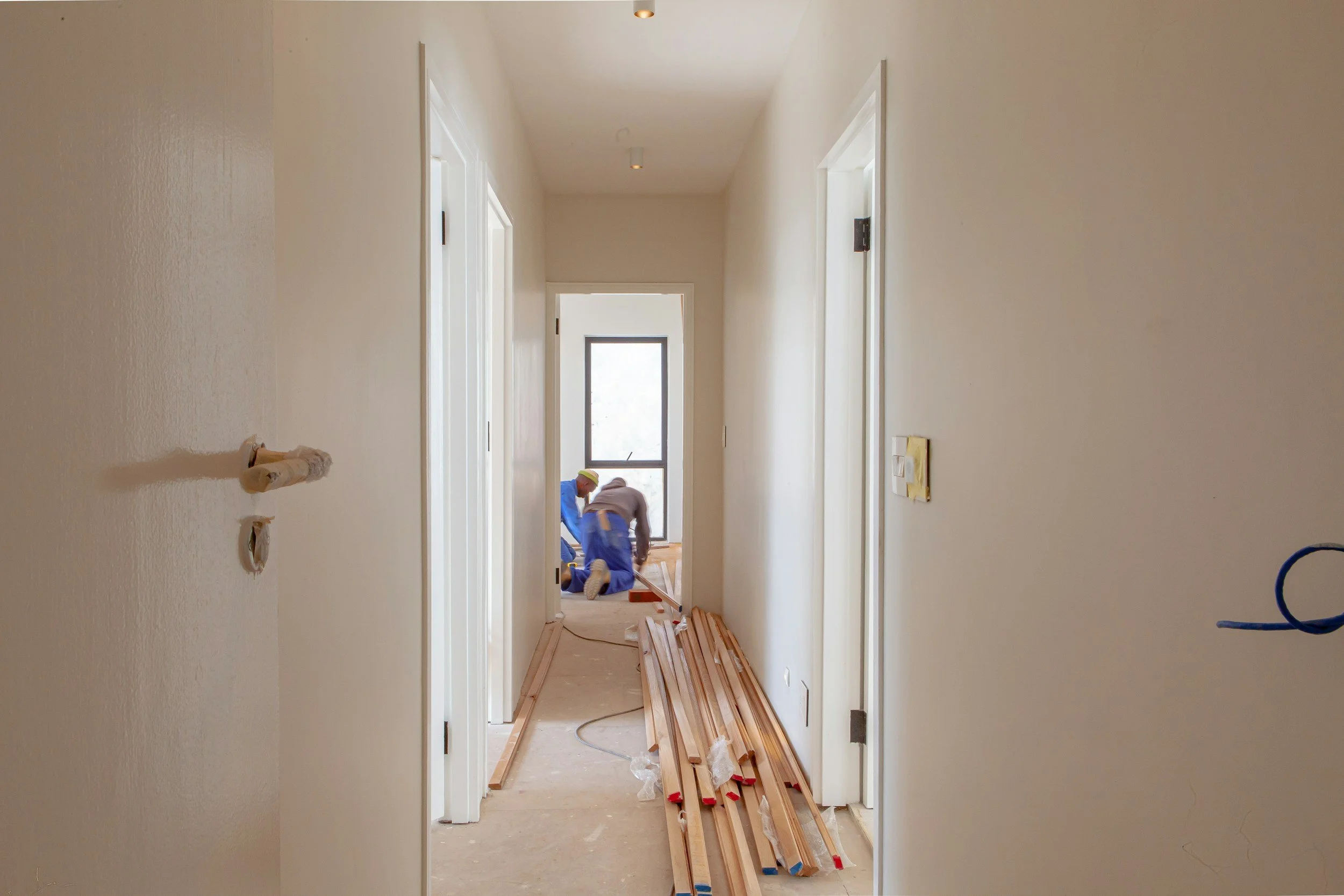 A hallway under construction with two construction workers kneeling near the end of the hallway, working on the floor, with wooden planks and construction tools scattered along the corridor.