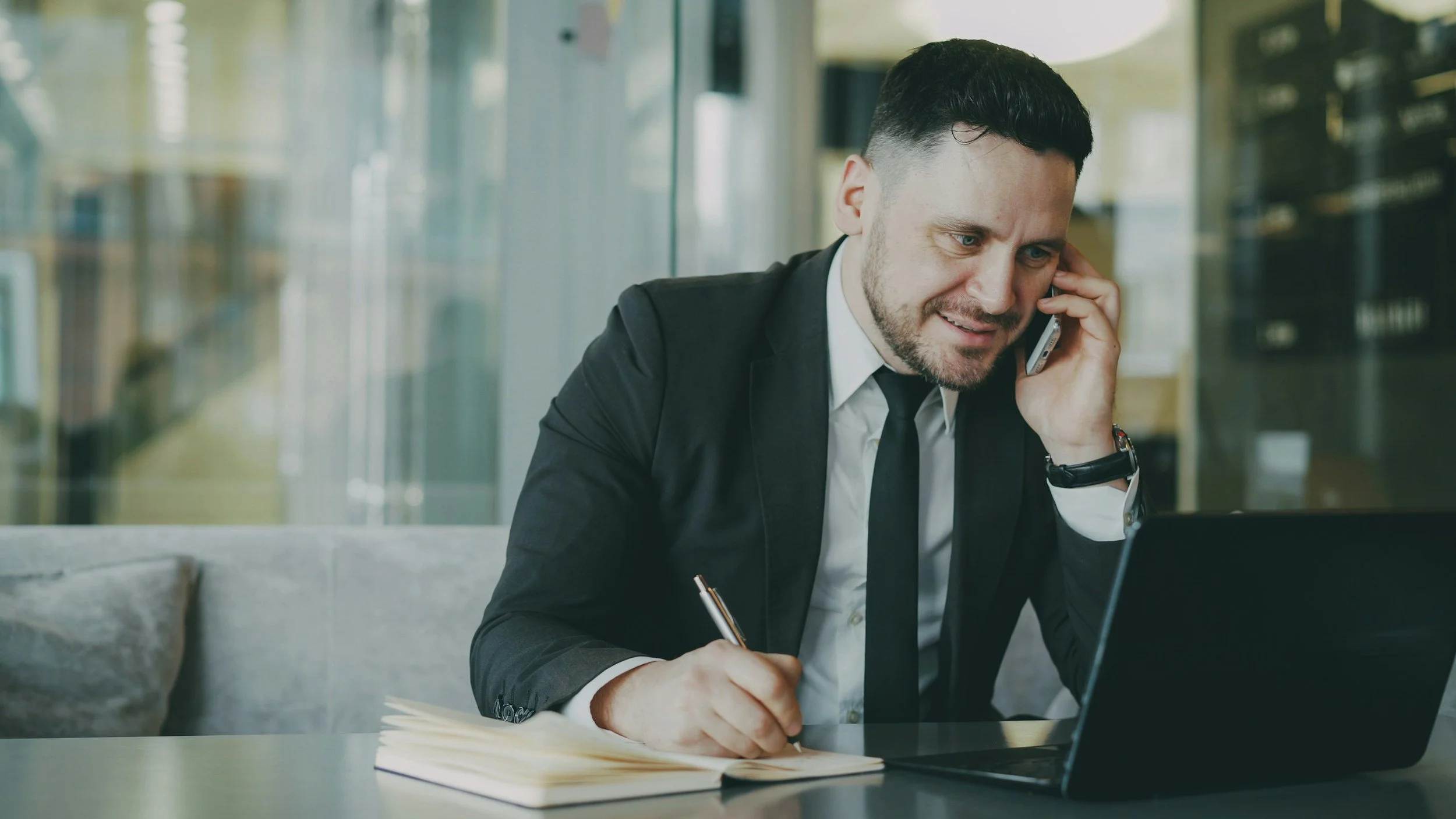 A man in a suit sitting at a desk in an office, talking on his cellphone, with a laptop and notepad in front of him.