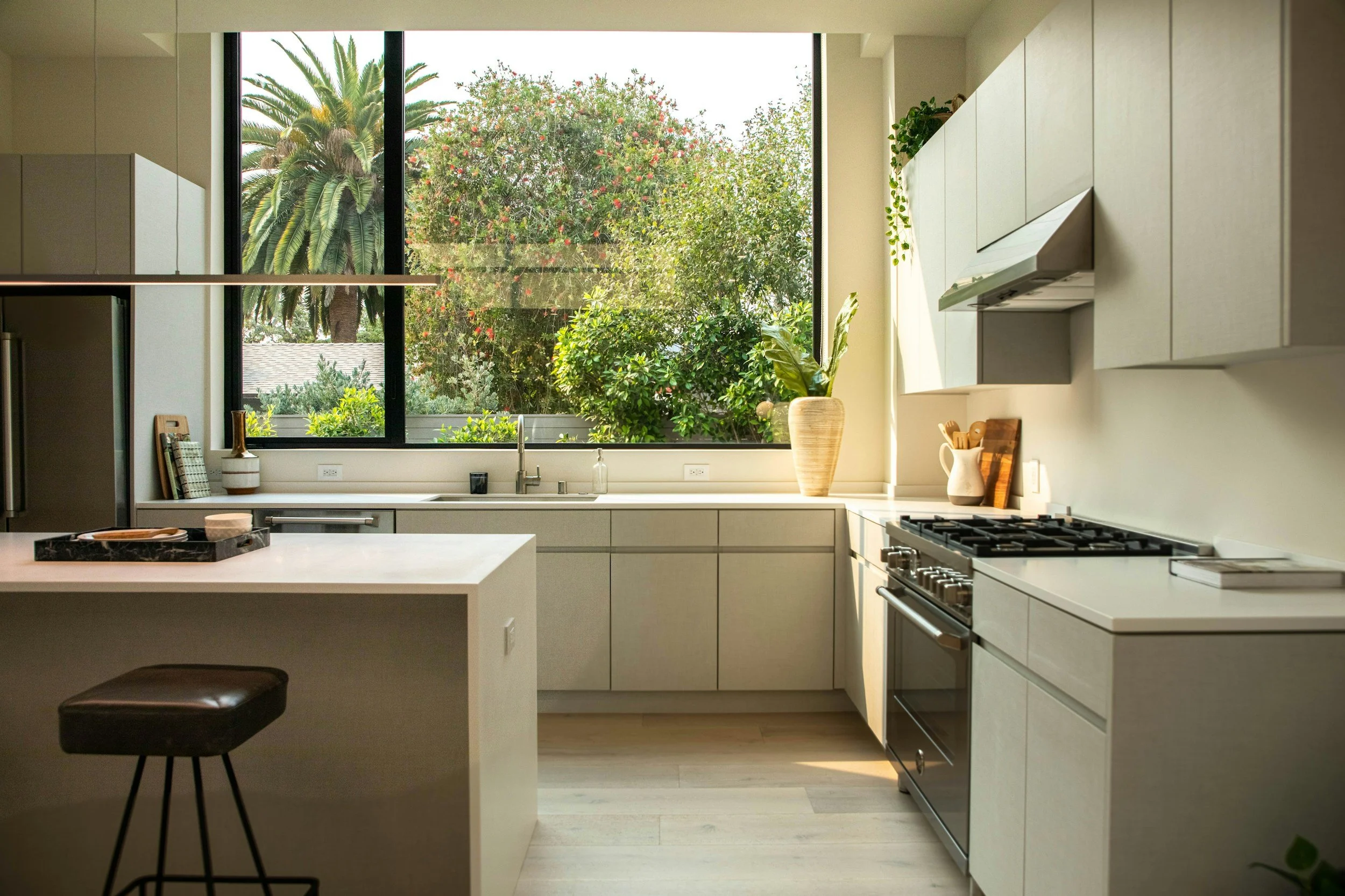 Modern kitchen with white cabinets, a large window with a view of trees and bushes outside, a kitchen island with a black stool, decorative items, and a stove.