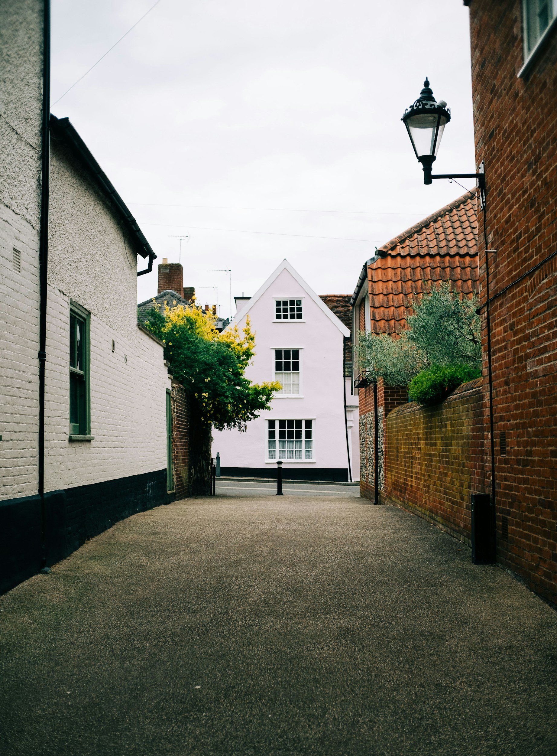 A quiet alleyway between buildings, with a dark asphalt surface, surrounded by brick and white painted walls, and trees with green and yellow leaves in the background. A vintage street lamp is attached to the right wall.