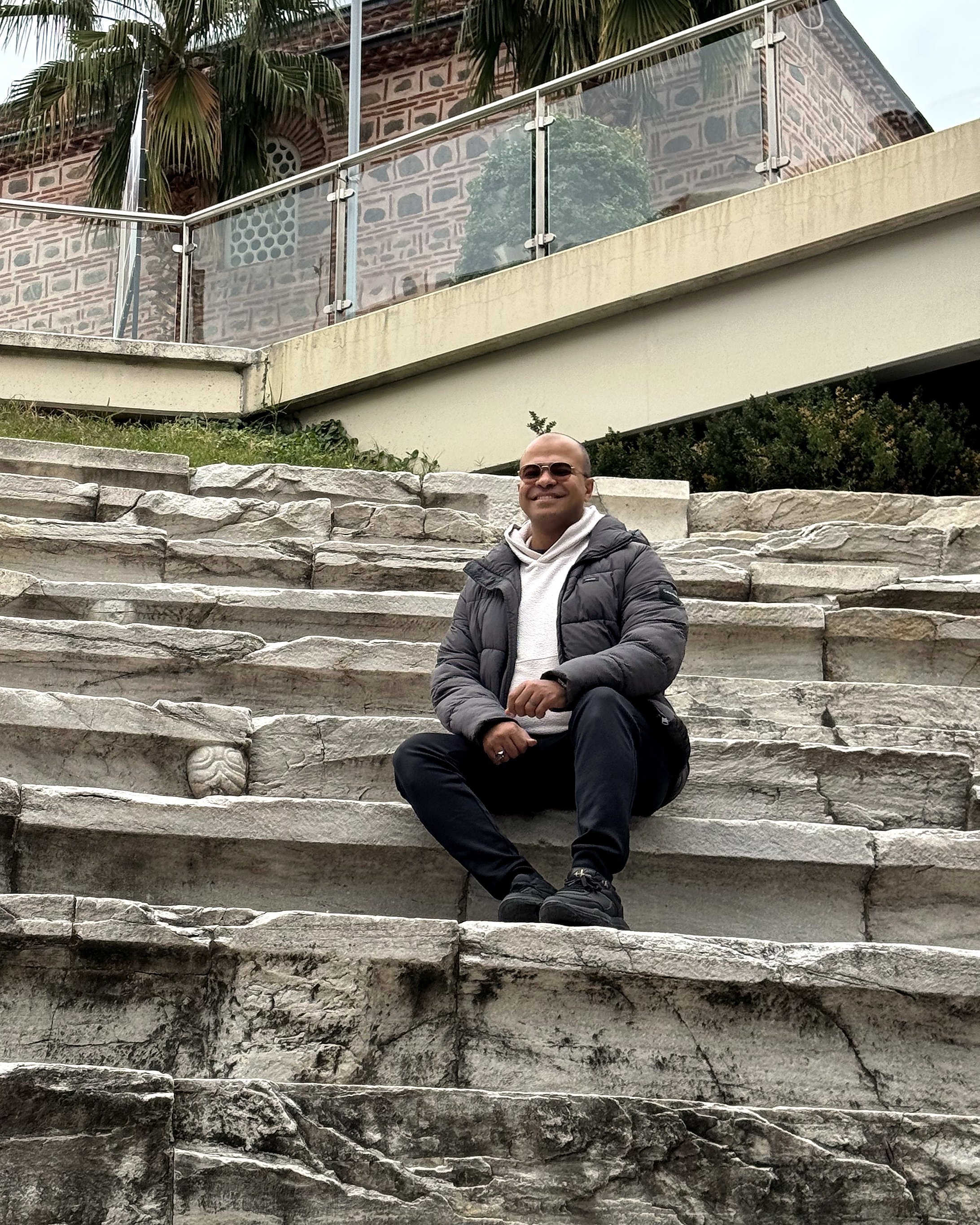 A man smiling and wearing sunglasses, a white hoodie, and a gray puffer jacket, sitting on stone steps outdoors.