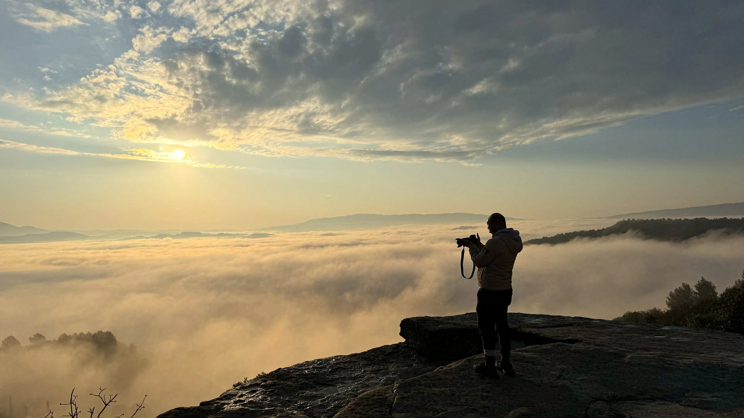 Person taking a photo of a sunrise over a foggy landscape from a rocky cliff.