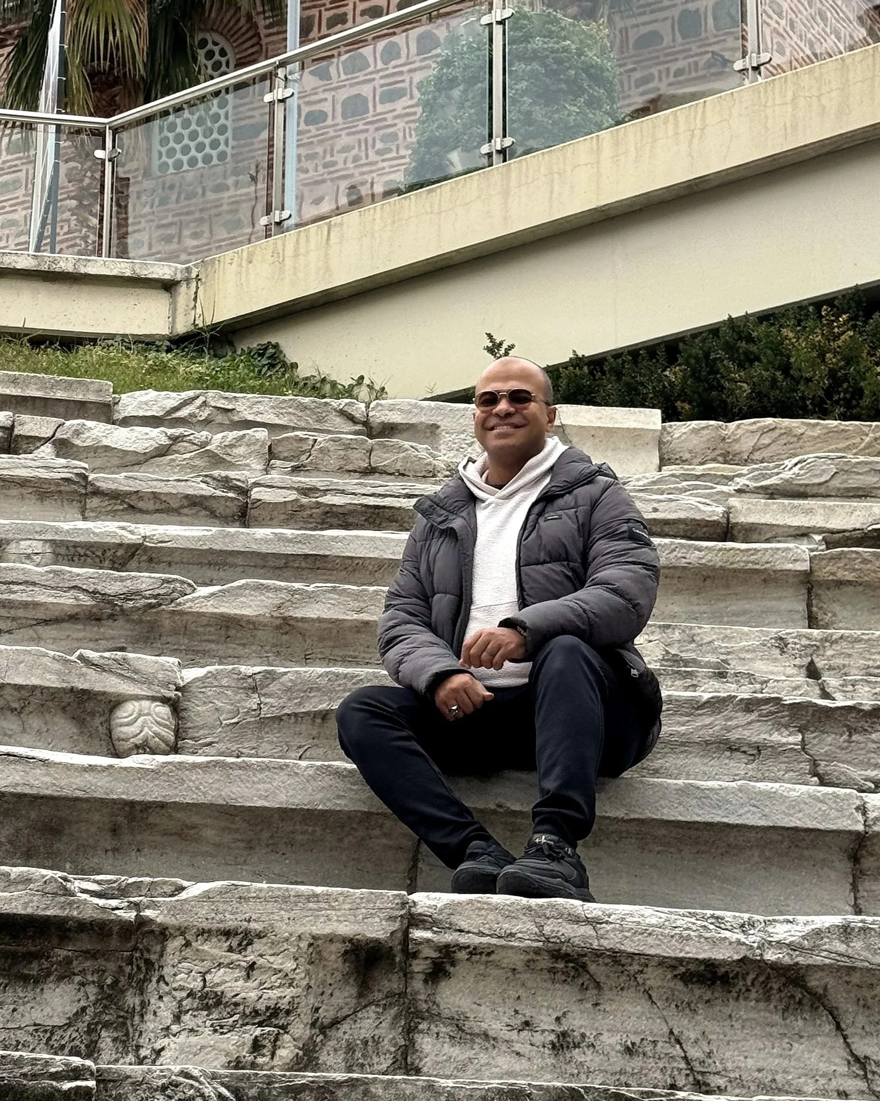 A man sitting on a stone staircase outdoors, smiling, wearing sunglasses, a gray jacket, white hoodie, black pants, and sneakers.