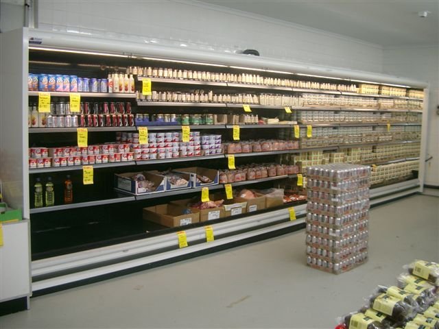 Grocery store aisle with shelves stocked with dairy products, beverages, and packaged foods, and a pallet of stacked canned goods in the aisle.