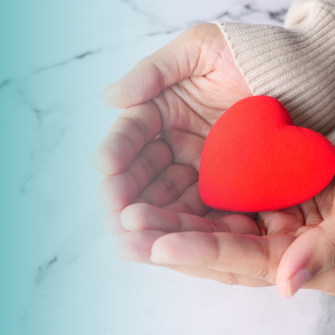 A person's hand holding a red foam heart with a beige sweater sleeve visible.