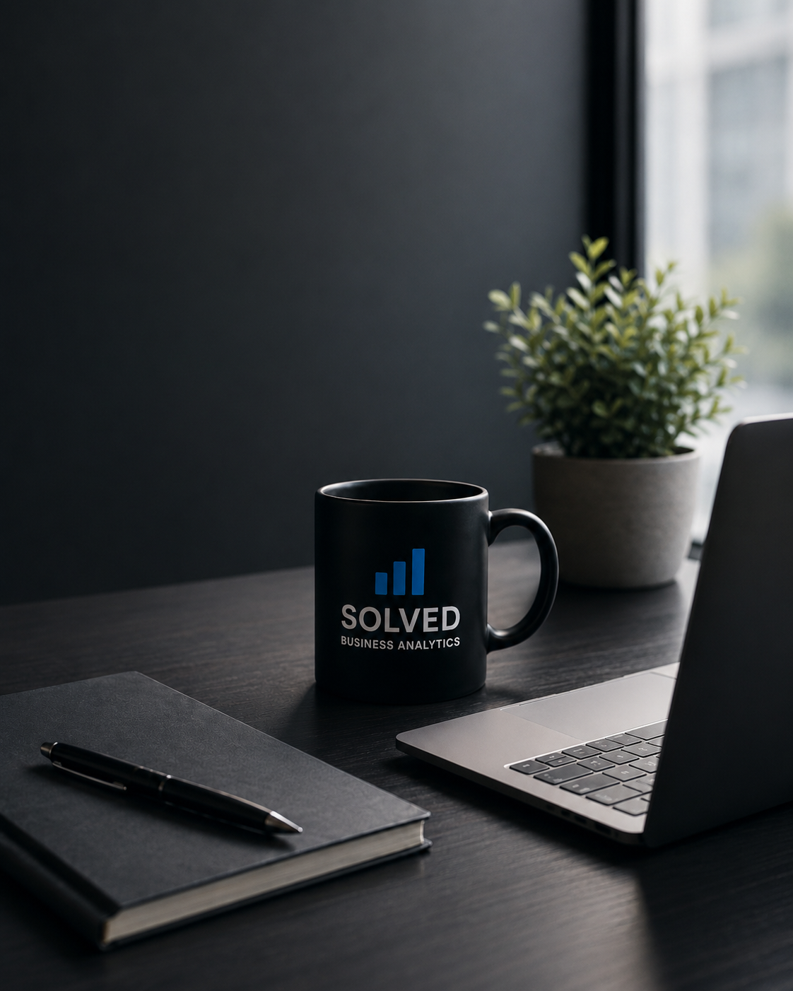 Black coffee mug with 'SOLVED Business Analytics' logo on a dark desk near a grey notebook and pen, with a laptop and potted plant in the background, illuminated by natural light from a window.