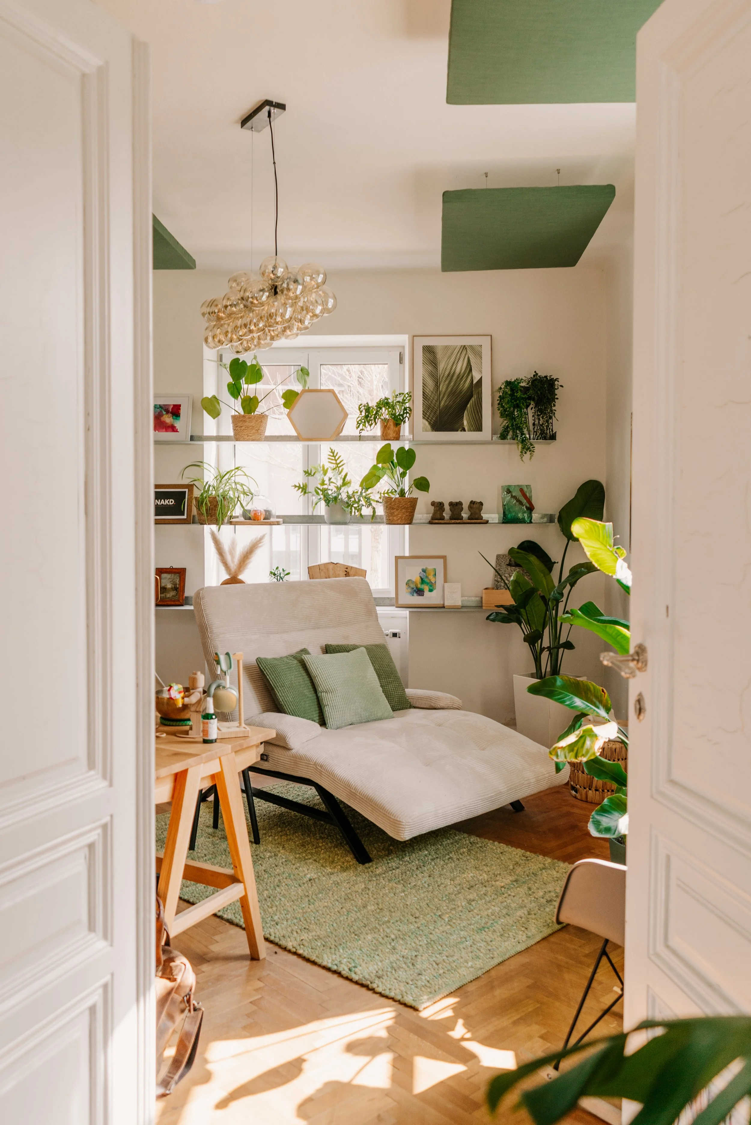 Ein hell eingerichtetes Wohnzimmer mit einem Sofa, grünen Kissen, Pflanzen, Wandbildern und einem Holzstuhl, gesehen durch eine offene Tür.