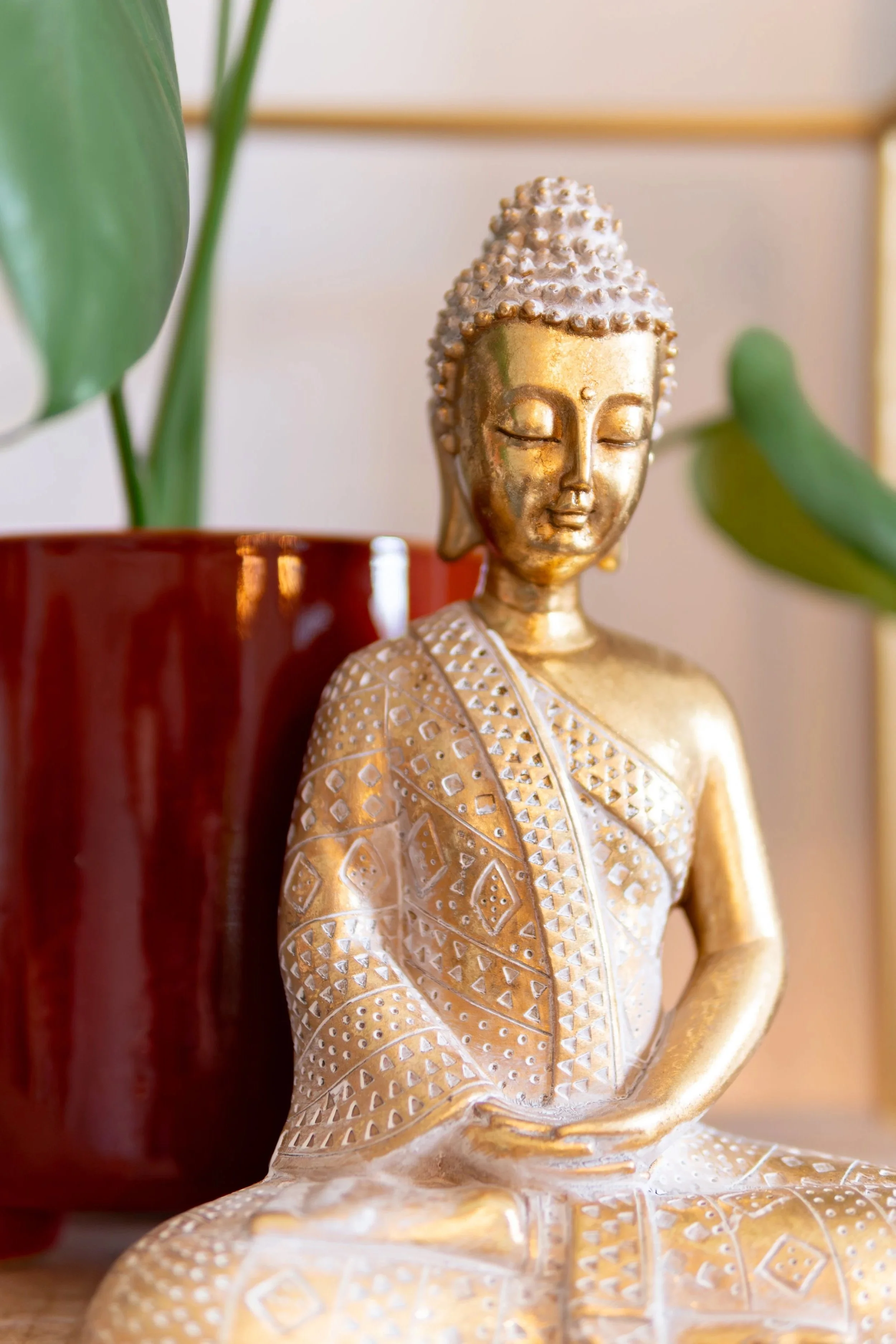 Goldene Buddha-Figur mit geschlossenen Augen neben eine grüne Pflanze in einem roten Topf