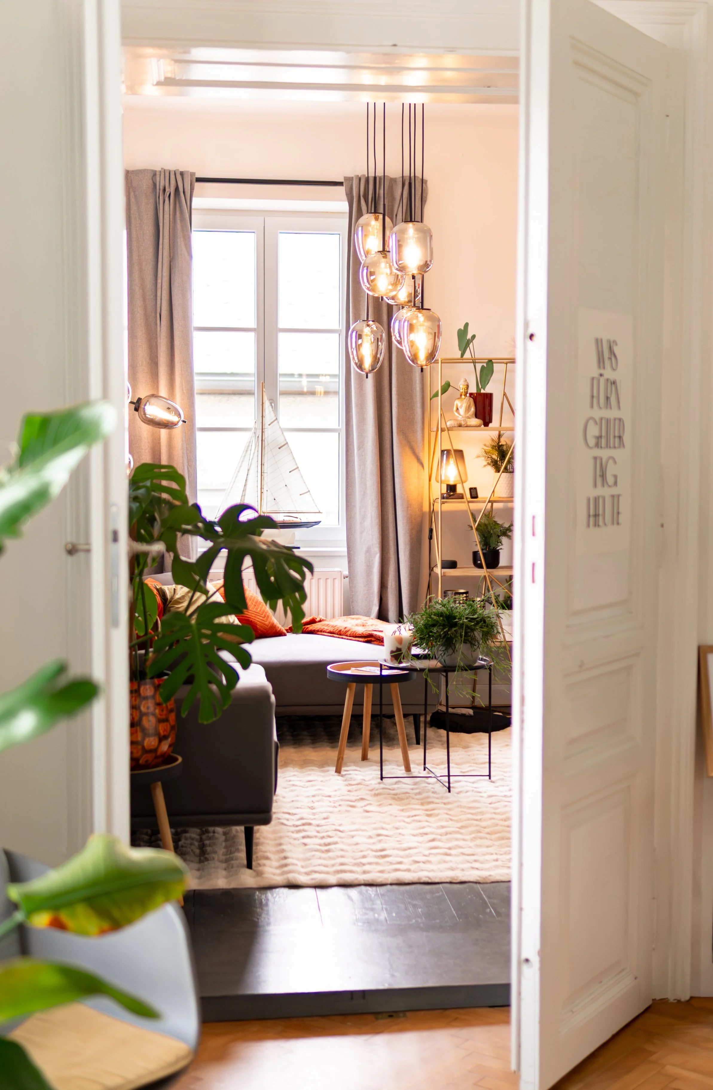 Einblick in ein Wohnzimmer durch eine offene Tür, mit Sofas, Couchtisch, mehreren Pflanzen, einer Stehlampe, Wanddekorationen und einem großen Fenster mit Vorhängen, beleuchtet durch Tageslicht und dekoriert mit modernen Lampen.