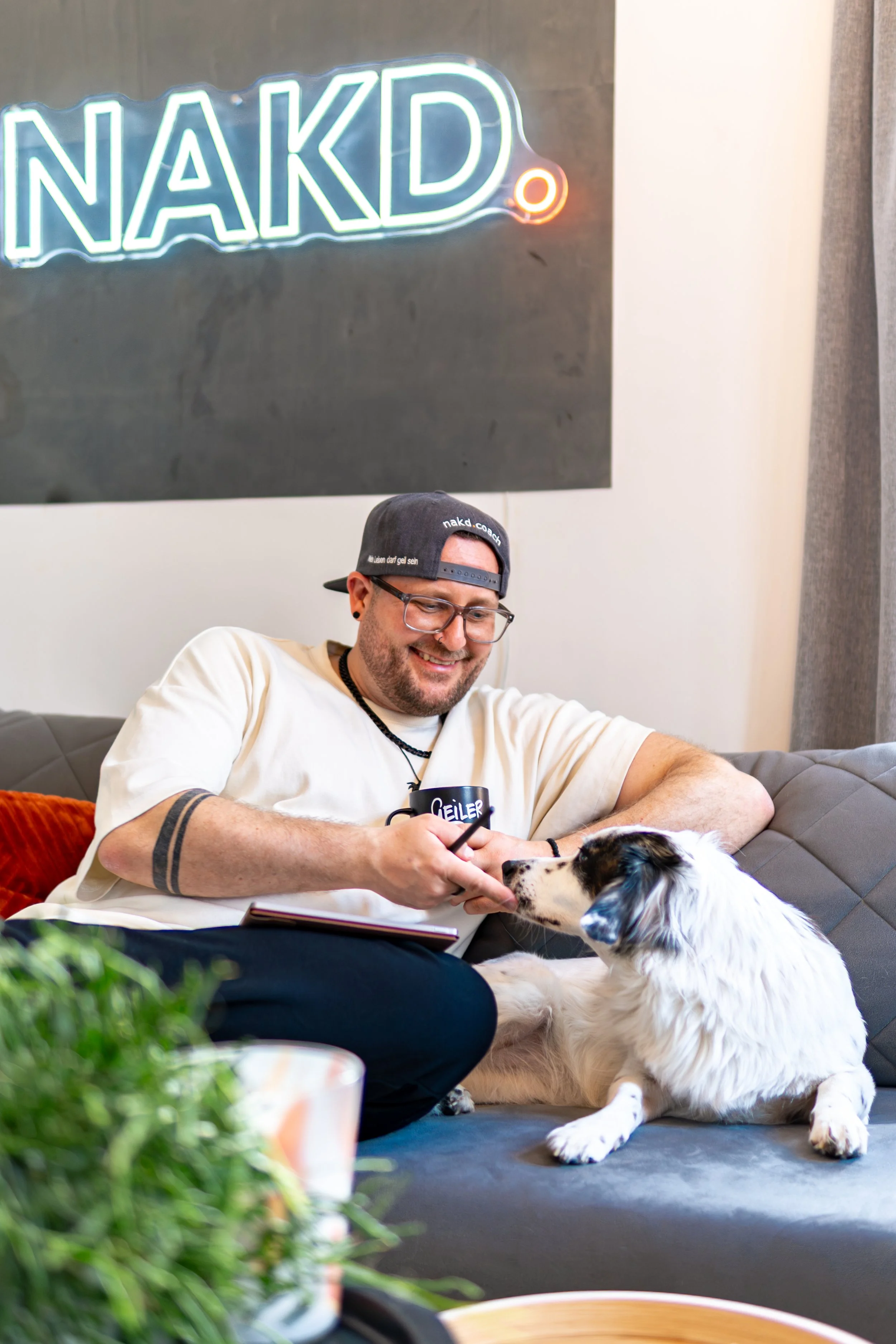 Ein Mann sitzt auf einem Sofa, lächelt und füttert einen Border Collie Hund mit seinem Finger, im Hintergrund eine leuchtende Neon-Schrift mit der Aufschrift "NAKD".
