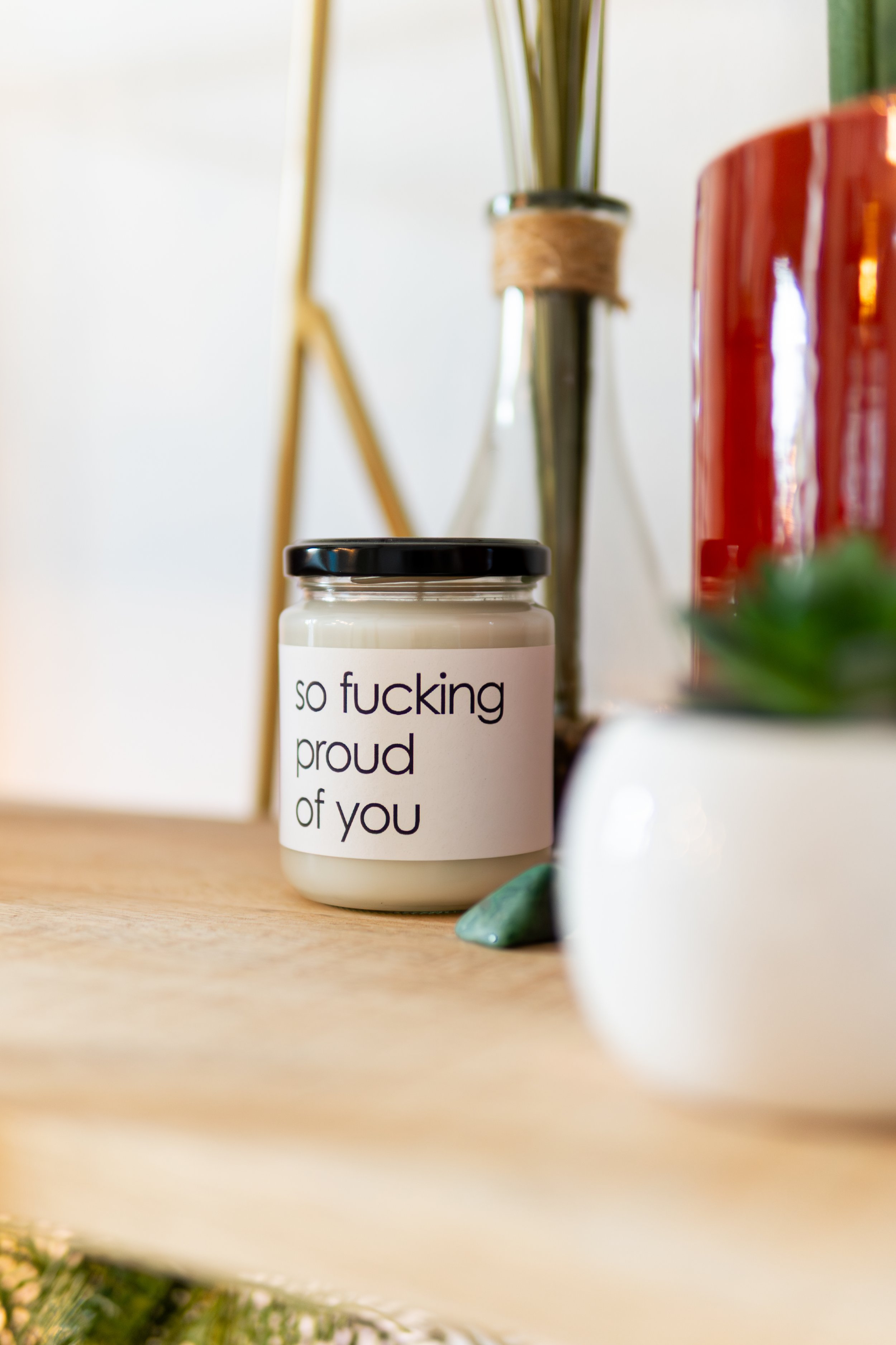 Ein großer Keramikbecher mit weißer Oberfläche und schwarzem Deckel, mit dem Text "so fucking proud of you". Der Becher steht auf einer Tischplatte, im Hintergrund Pflanzen und Dekorationsgegenstände, die leicht unscharf sind.