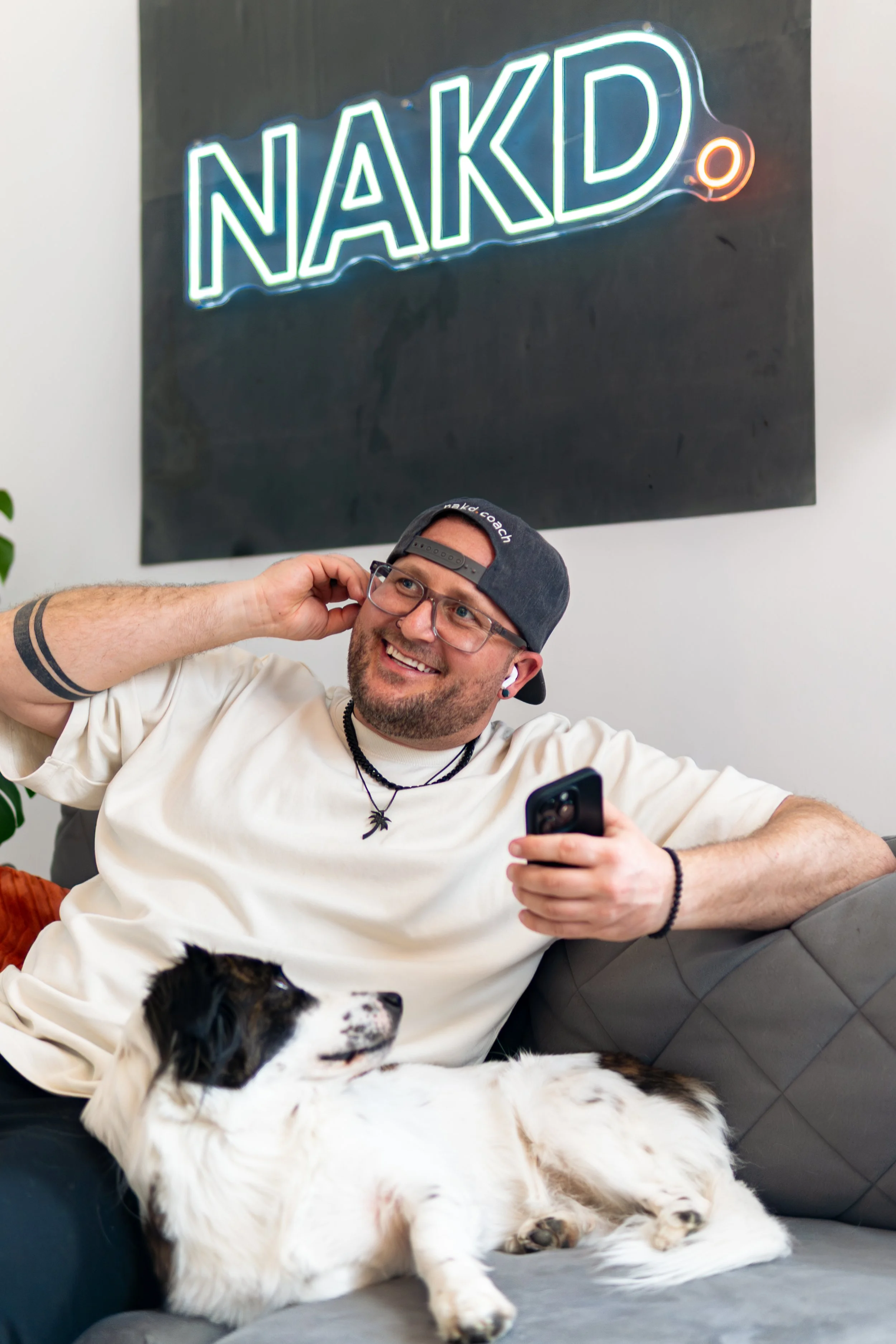 Ein glücklicher Mann mit Brille, rotem Hut und einem Hund auf dem Schoß, der ein Telefon hält, sitzt auf einem grauen Sofa. Im Hintergrund befindet sich eine schwarze Wand mit einem neonblauen Schriftzug "NAKD" und einem kleinen roten Punkt.