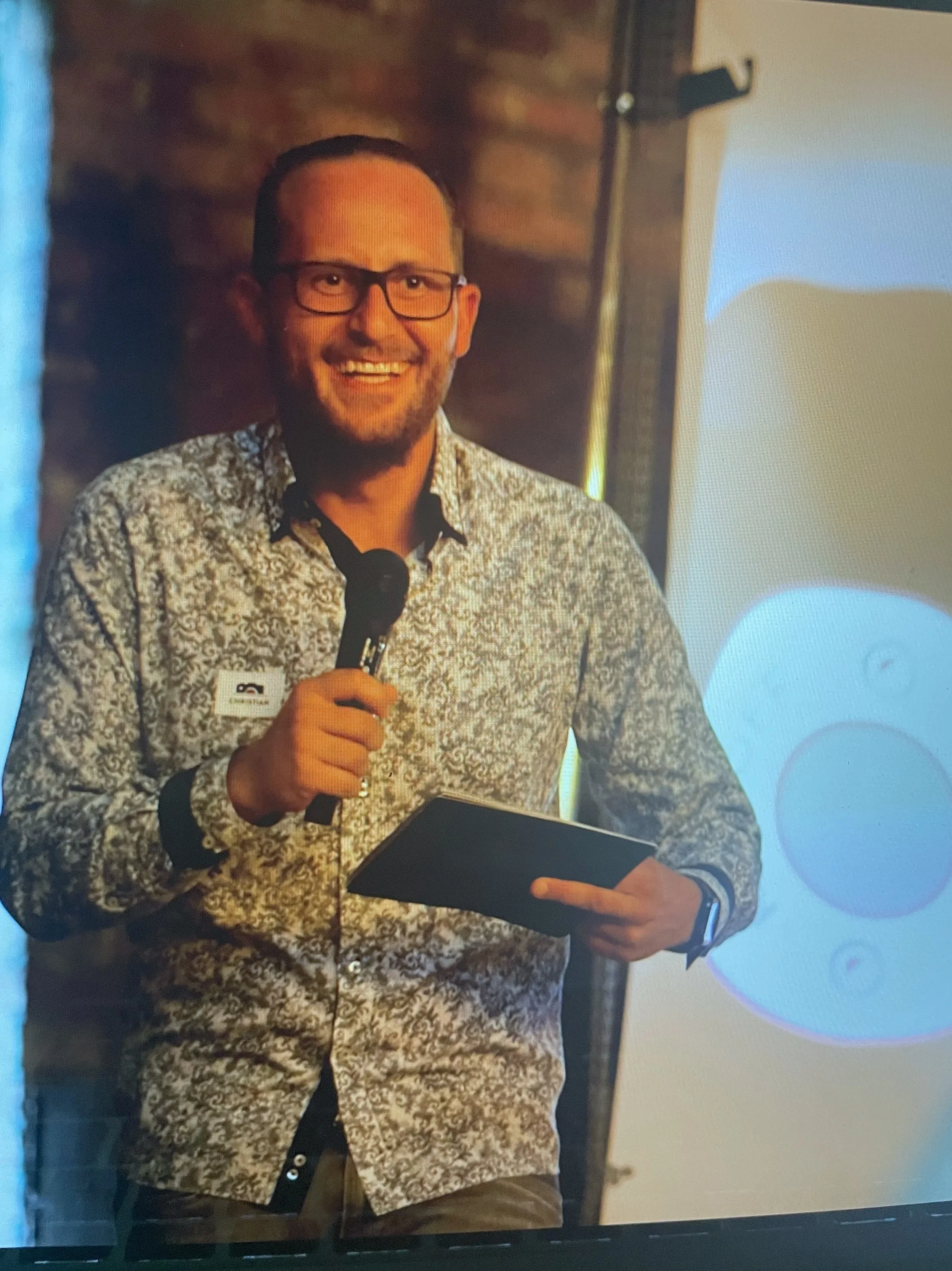 Ein Mann mit Brille, beerbtem grauem Hemd mit schwarzer Musterung, hält ein Tablet und spricht in ein Mikrofon, vermutlich bei einer Präsentation oder Veranstaltung.