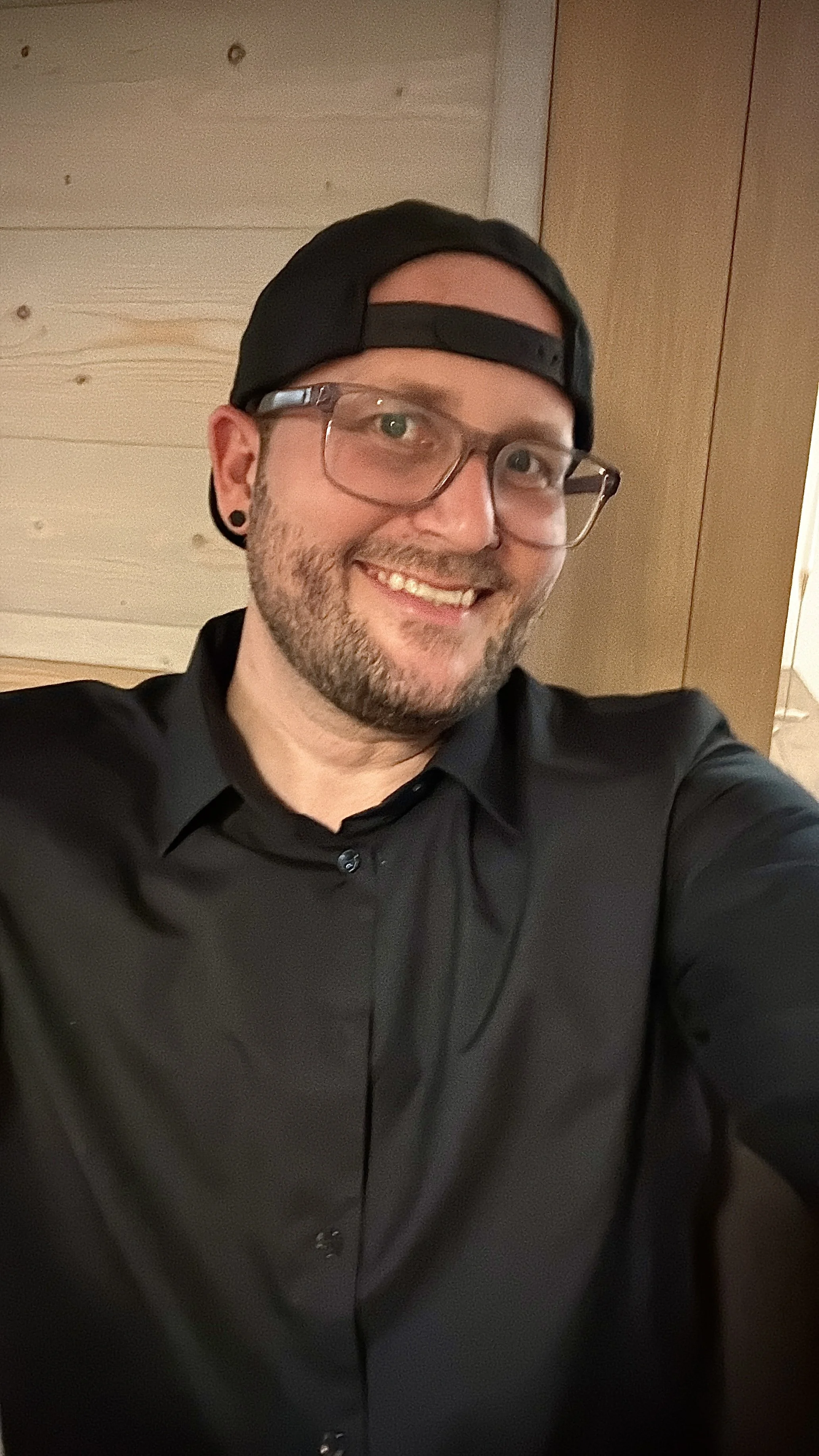 Ein lachender Mann mit Brille, schwarzer Kappe, schwarzen Hemd, im Hintergrund eine Holzwand.