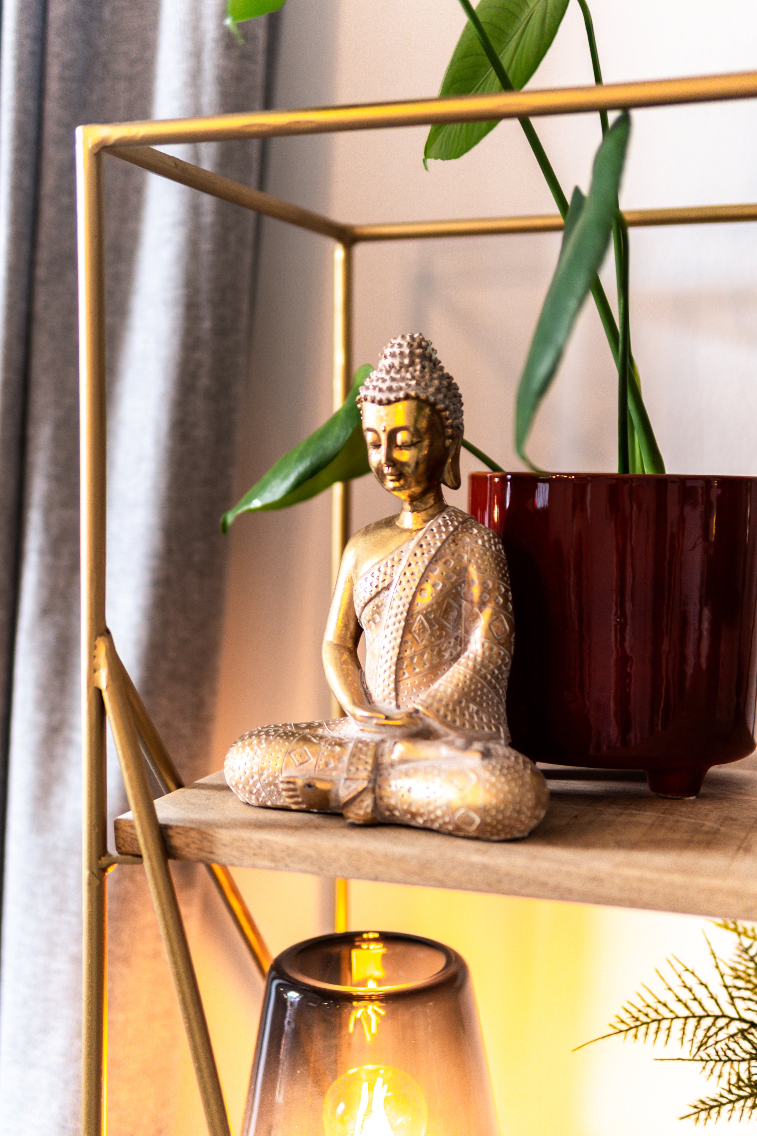 Ein goldener Buddha-Statue auf einem Holzregal neben grünen Pflanzen in einer roten Topf. Im Hintergrund eine graue Gardine und eine leuchtende Glühbirne in einer schwarzen Lampe.