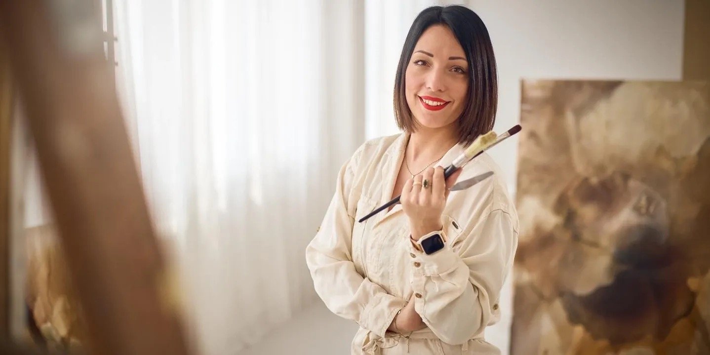 Frau mit dunklen Haaren, rotem Lippenstift und einem weißen Anzug hält Pinsel und Palette, steht vor einem Gemälde in einem Atelier.