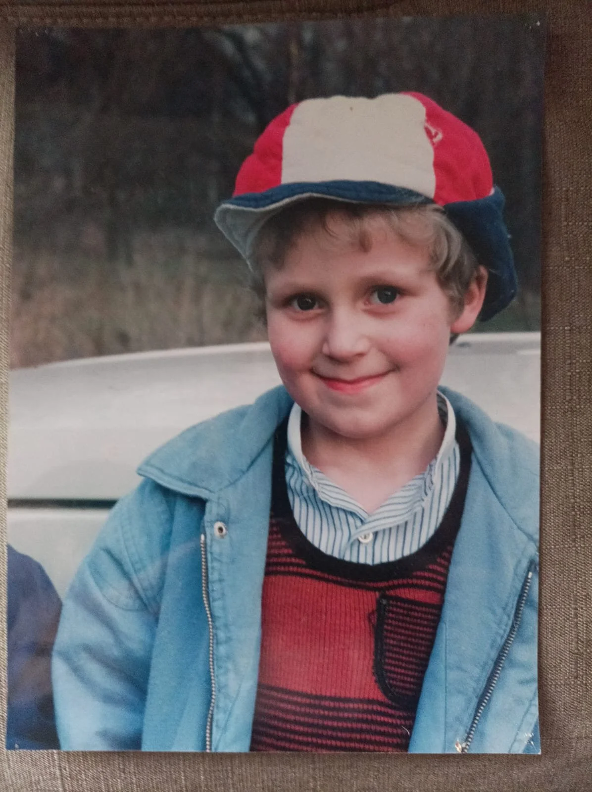 Ein Junge mit blauen Augen, hellbraunen Haaren und einem Lächeln, trägt eine rote, weiße und blaue Mütze, einen blauen Denim-Jacken, ein gestreiftes Hemd und einen roten Pullover mit schwarzen Linien, steht vor einem Auto