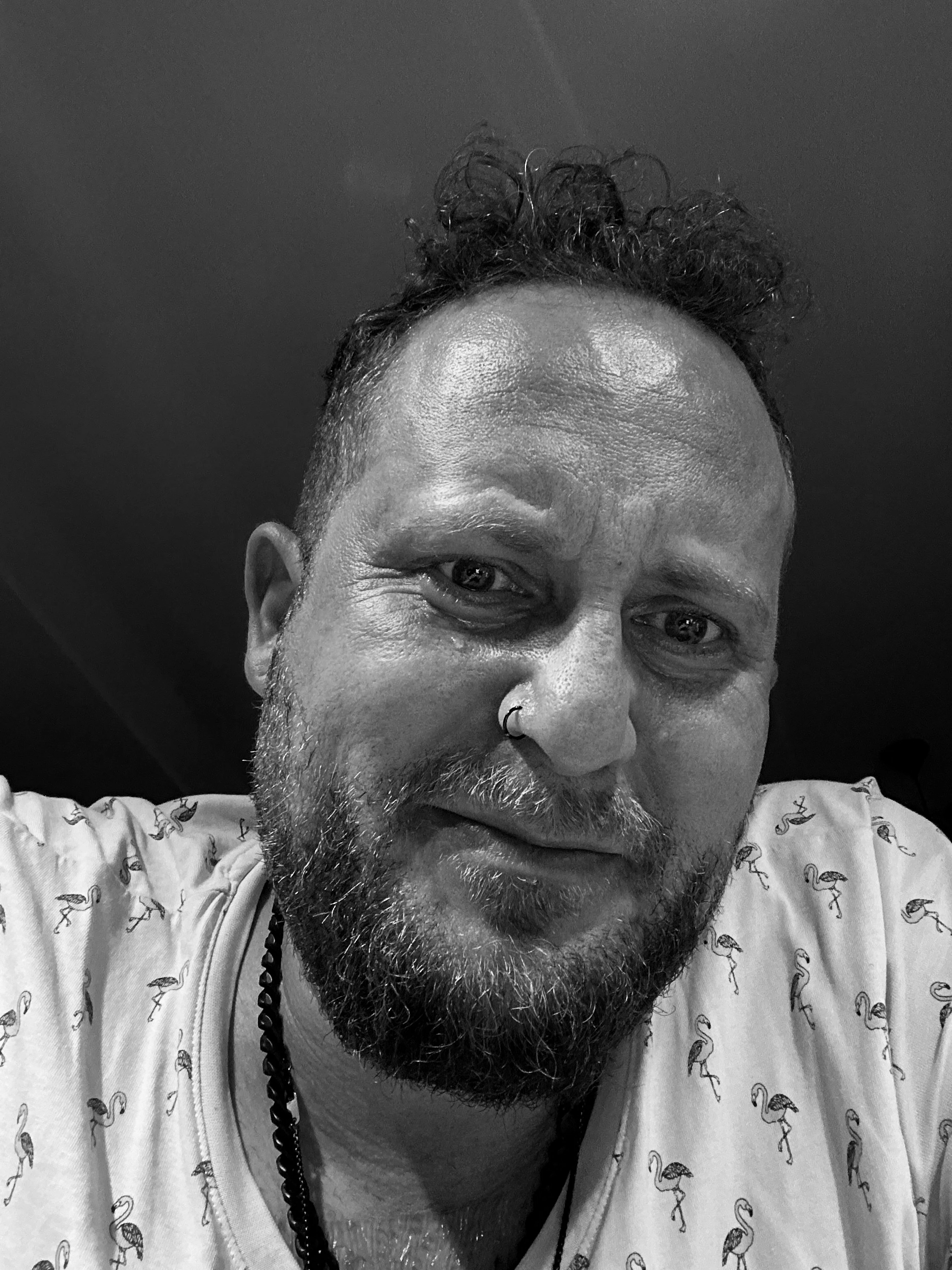 Schwarz-weiß-Foto eines Mannes mit beards, kurzer Frisur und Nasenpiercing, trägt ein T-Shirt mit Flamingo-Muster und eine Halskette, lächelt leicht.