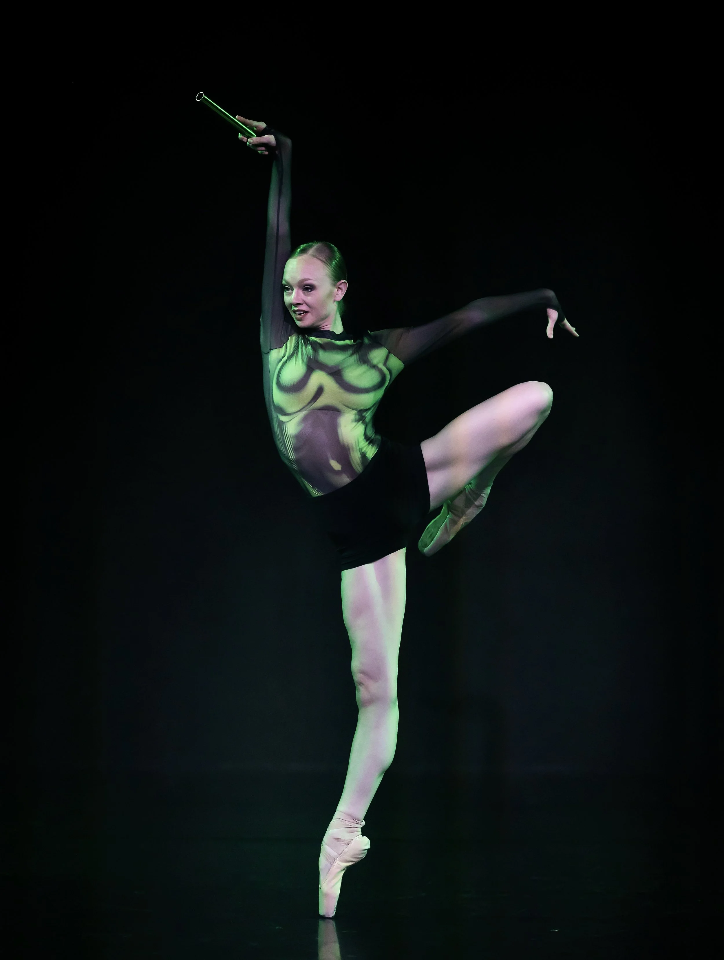 Balletttänzerin in einer Pose, trägt einen schwarzen Blick mit grüner Beleuchtung, auf dunklem Hintergrund.