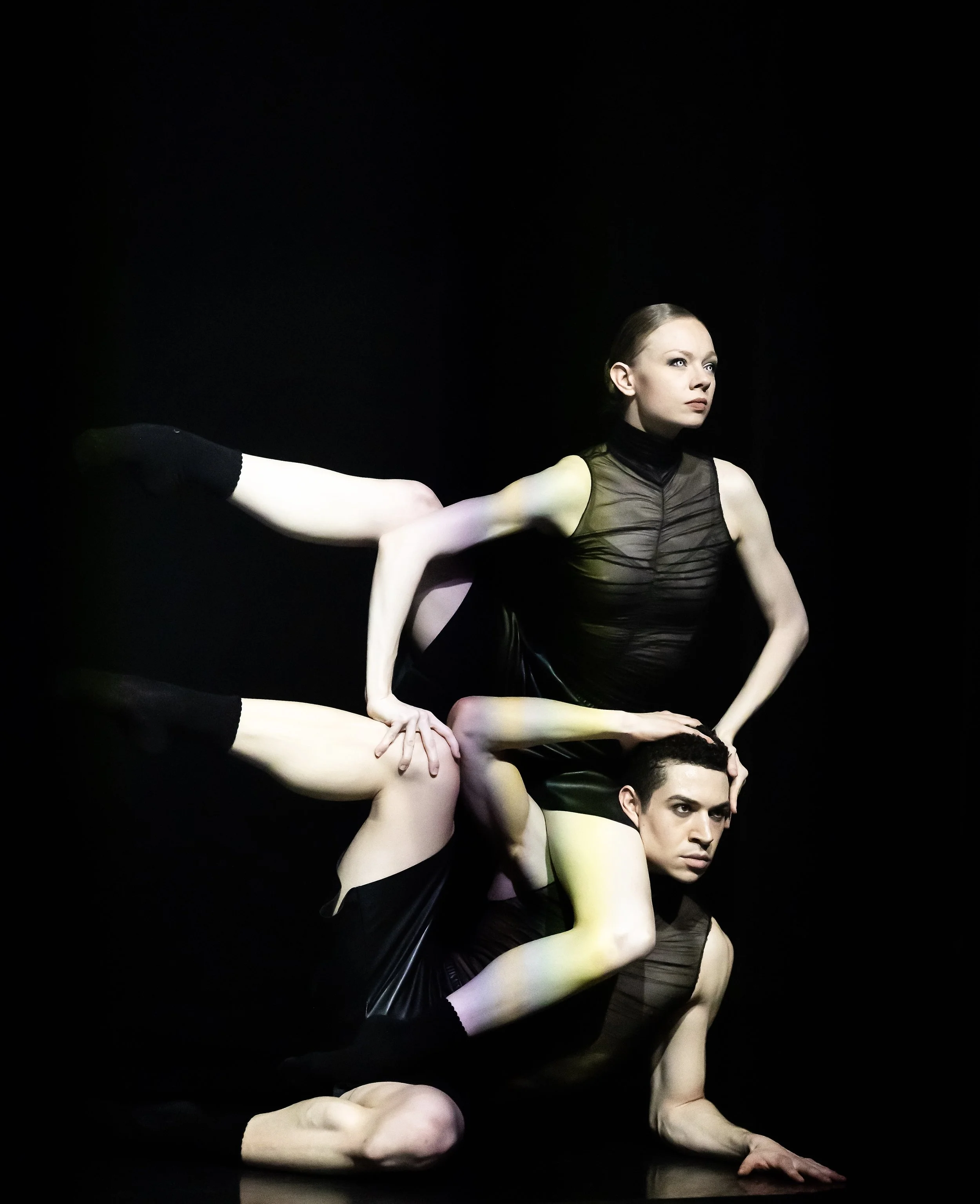 Mehrere Tänzer in schwarzen Outfits in einer Performance, die auf einem schwarzen Hintergrund auftreten, mit fokus auf ihren Körperhaltungen und Bewegungen.