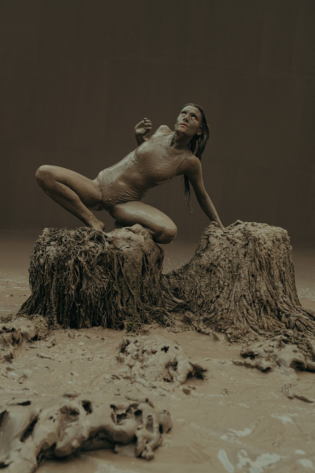 Kunstinstallation einer hautfarbenen weiblichen Figur in einer gebückten Haltung auf einem Baumstumpf, umgeben von Tierköpfen, in einer künstlerischen Darstellung mit puristischem Hintergrund.