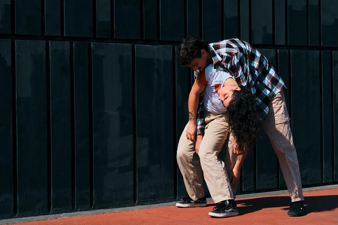 Zwei Jugendliche, die sich an einem schwarzen Wand mit einer roten Bodenfläche befinden, eine Person trägt eine kariertes Hemd und die andere eine weiße T-Shirt.