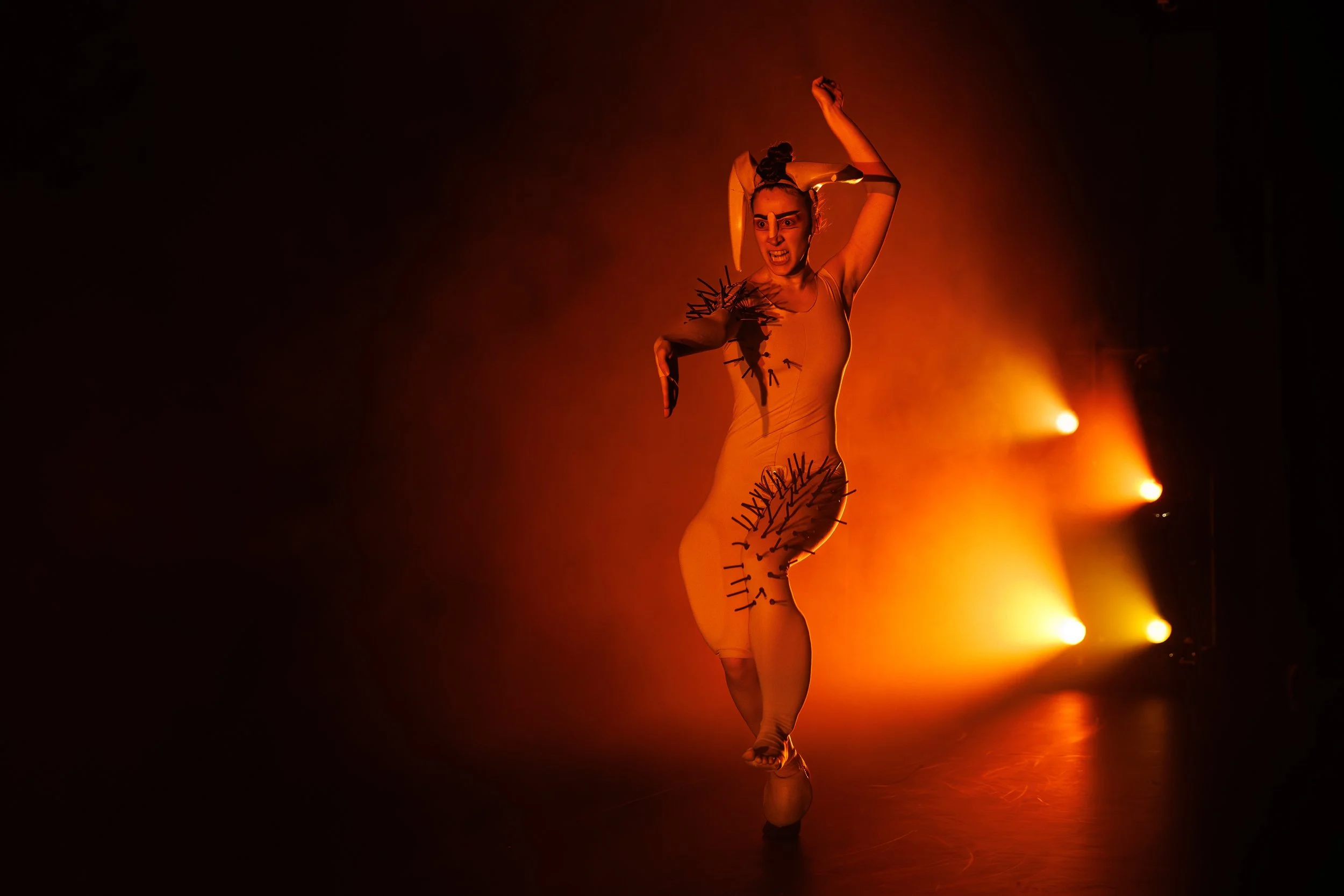 Tänzerin während einer Performance in einem orange beleuchteten Raum, trägt ein Kostüm mit schwarzen Stichwörtern und Papiertüten an den Armen, die nervös wirkt.