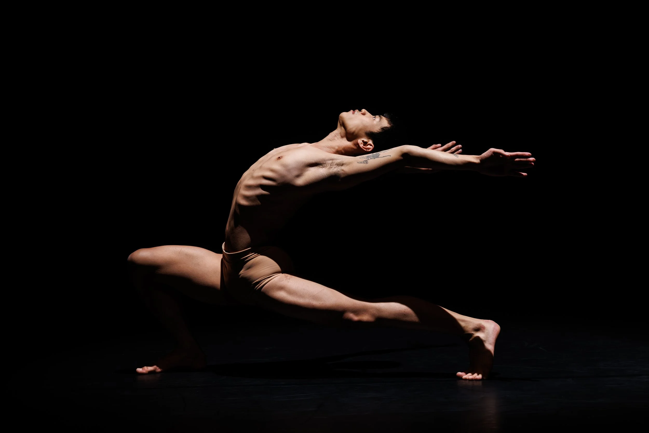 Ein männlicher Tänzer in einer komplexen Yoga- oder Tanzpose auf einer dunklen Bühne, nur durch ein Licht beleuchtet.