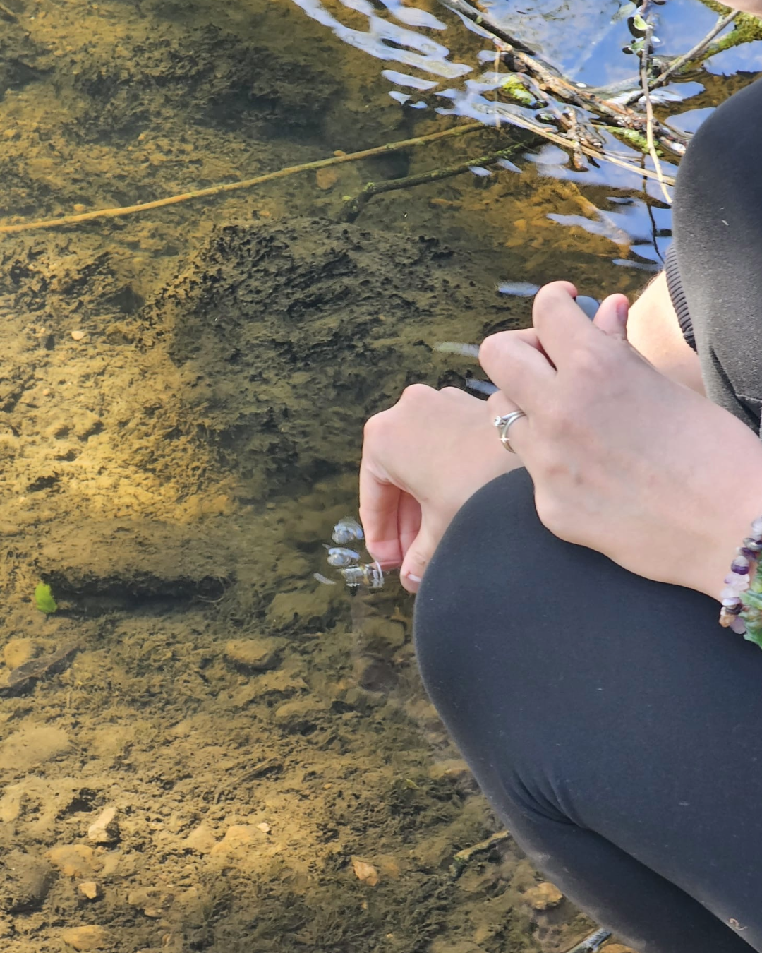 Person sitting by a shallow water stream, touching the water with their fingers, wearing a ring and jewelry, with the view of the sandy and muddy bottom of the stream.