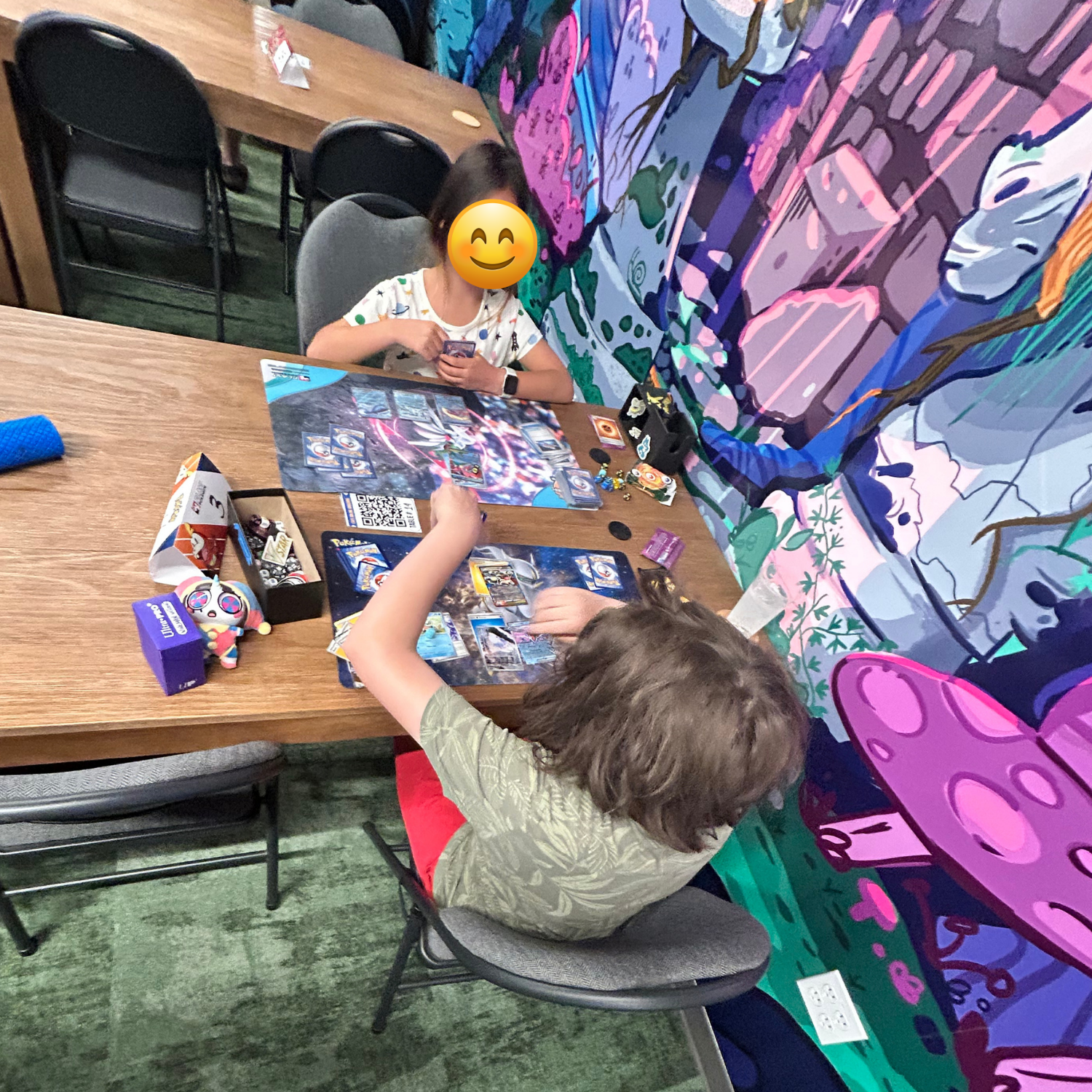 Two children playing Pokémon at  table, with various game accessories and cards spread out. One child is smiling with an emoji face, while the other is focused on the game. The background features a colorful mural with pink, purple, and green hues.