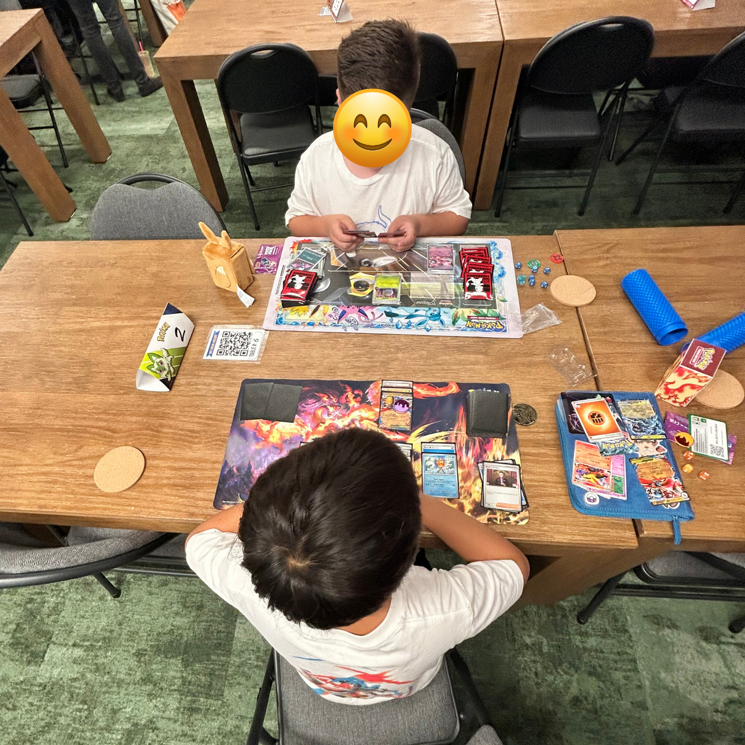 Two children playing a Pokémon trading card game at a wooden table in an indoor setting, with board game accessories and cards scattered on the table.