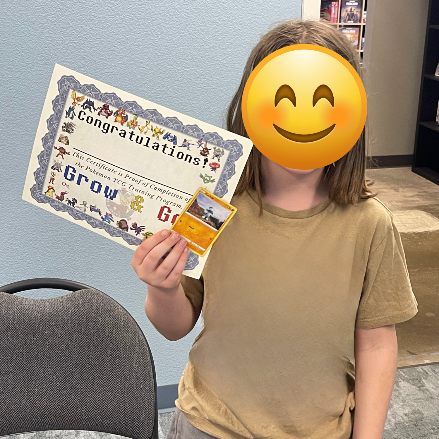A young girl is holding a Pokémon card and a certificate of completion for the Pokémon TCG training program, while smiling beneath a smiley face emoji. She is wearing a brown T-shirt and standing indoors with a blue wall and bookshelves in the background.