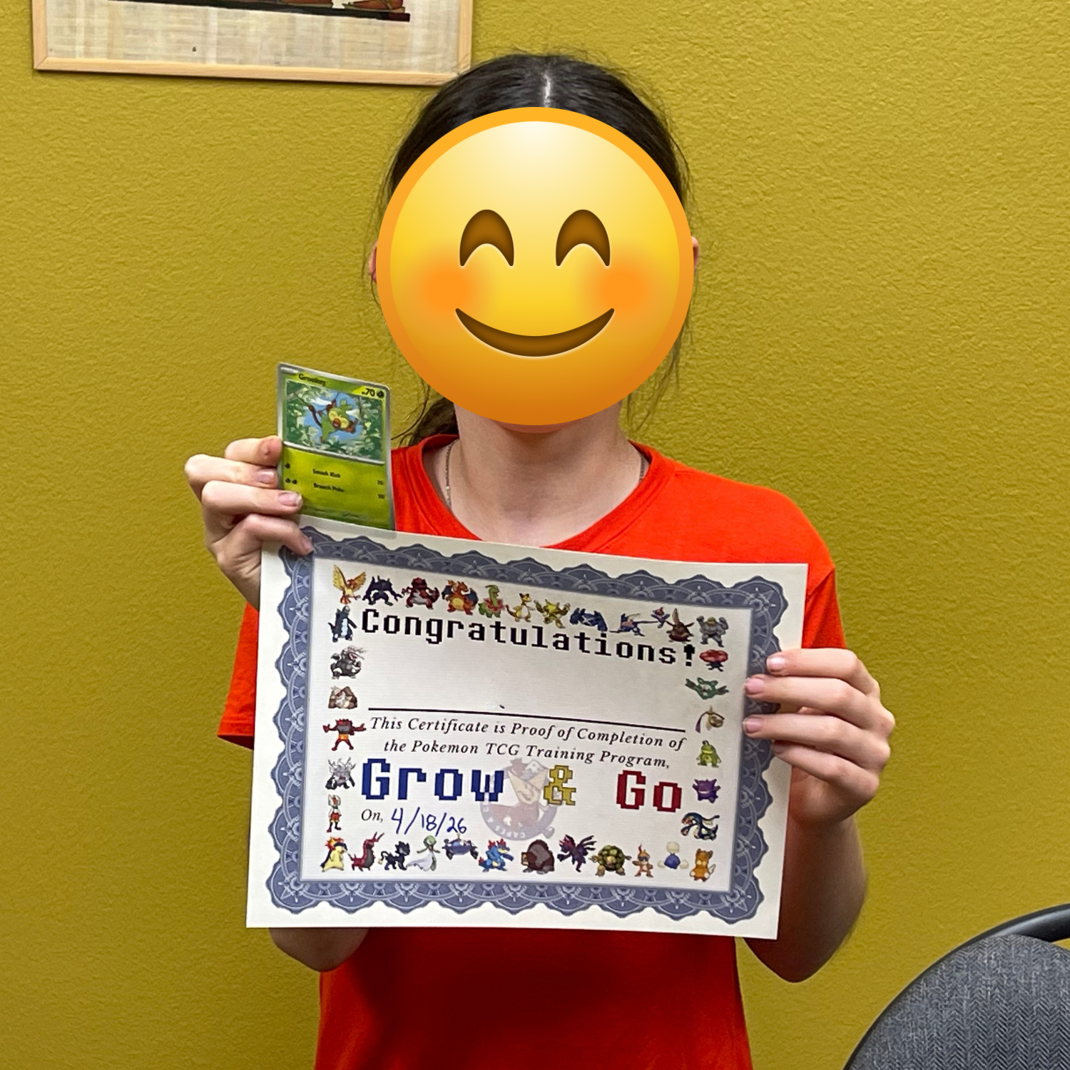 A person holding a Pokémon-themed graduation certificate and a Pokémon trading card in front of a yellow wall.
