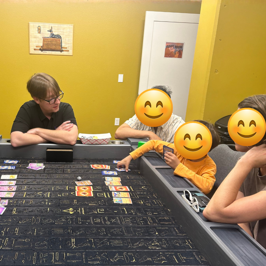 Four people with emoji faces sit around a table playing Pokémon card game.