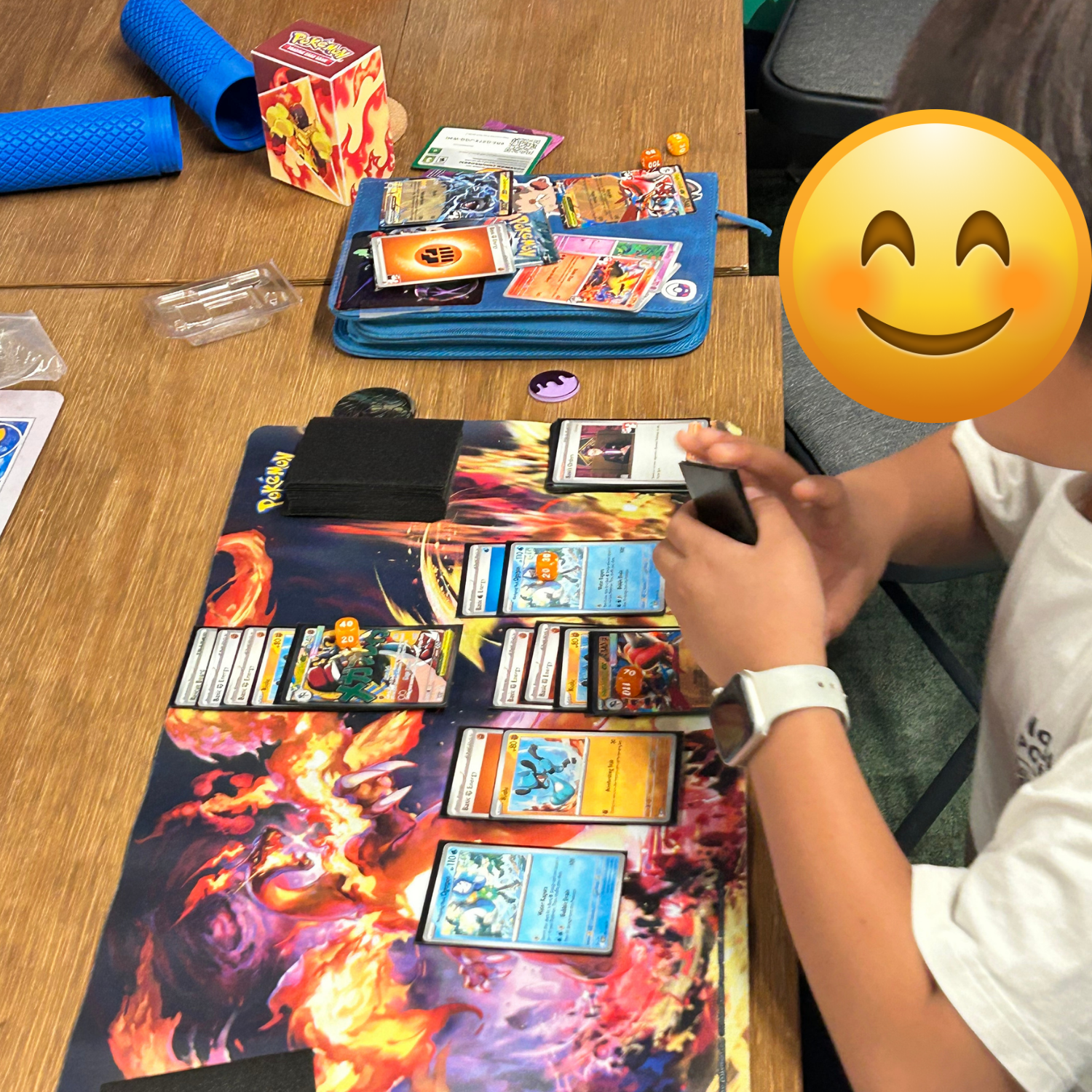A person playing a card game on a Pokémon-themed playmat, with Pokémon cards laid out on the table, a blue playmat rolled up at the side, and a Pokémon booster box and dice on the table.
