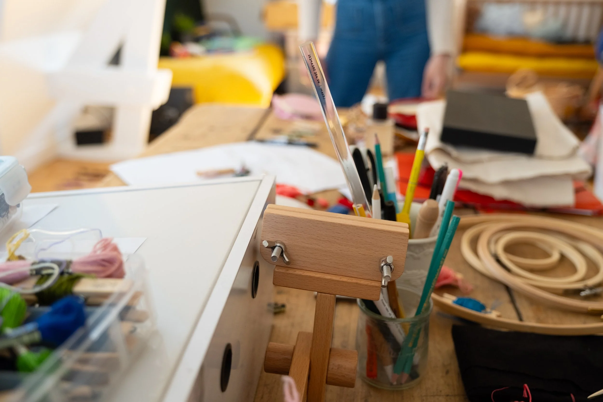 A cluttered workspace with various art supplies including colored pencils, pens, and sewing tools. A person wearing jeans is visible in the background, and the table is filled with paper, fabric, and other creative materials.