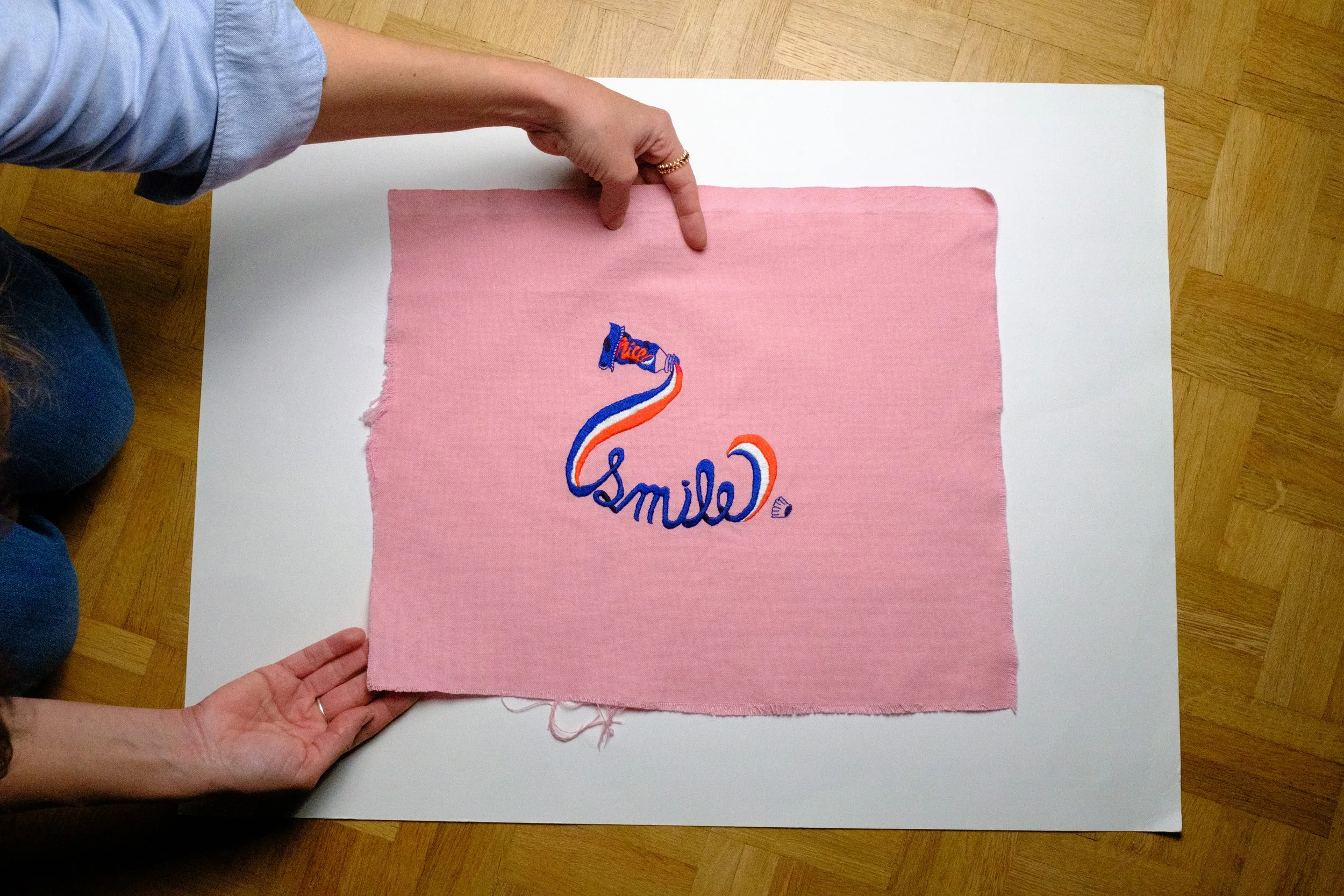 Two hands hold a pink fabric with embroidered blue, red, and orange text and design, placed on a white surface on a wooden floor. The fabric has the word 'Smile' embroidered on it.