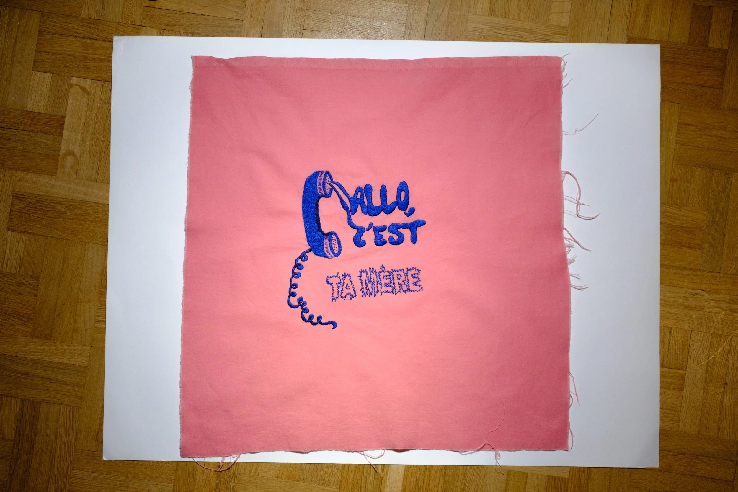 Pink fabric with an embroidery that features a blue telephone receiver and the French words "Allo, c'est ta mère."