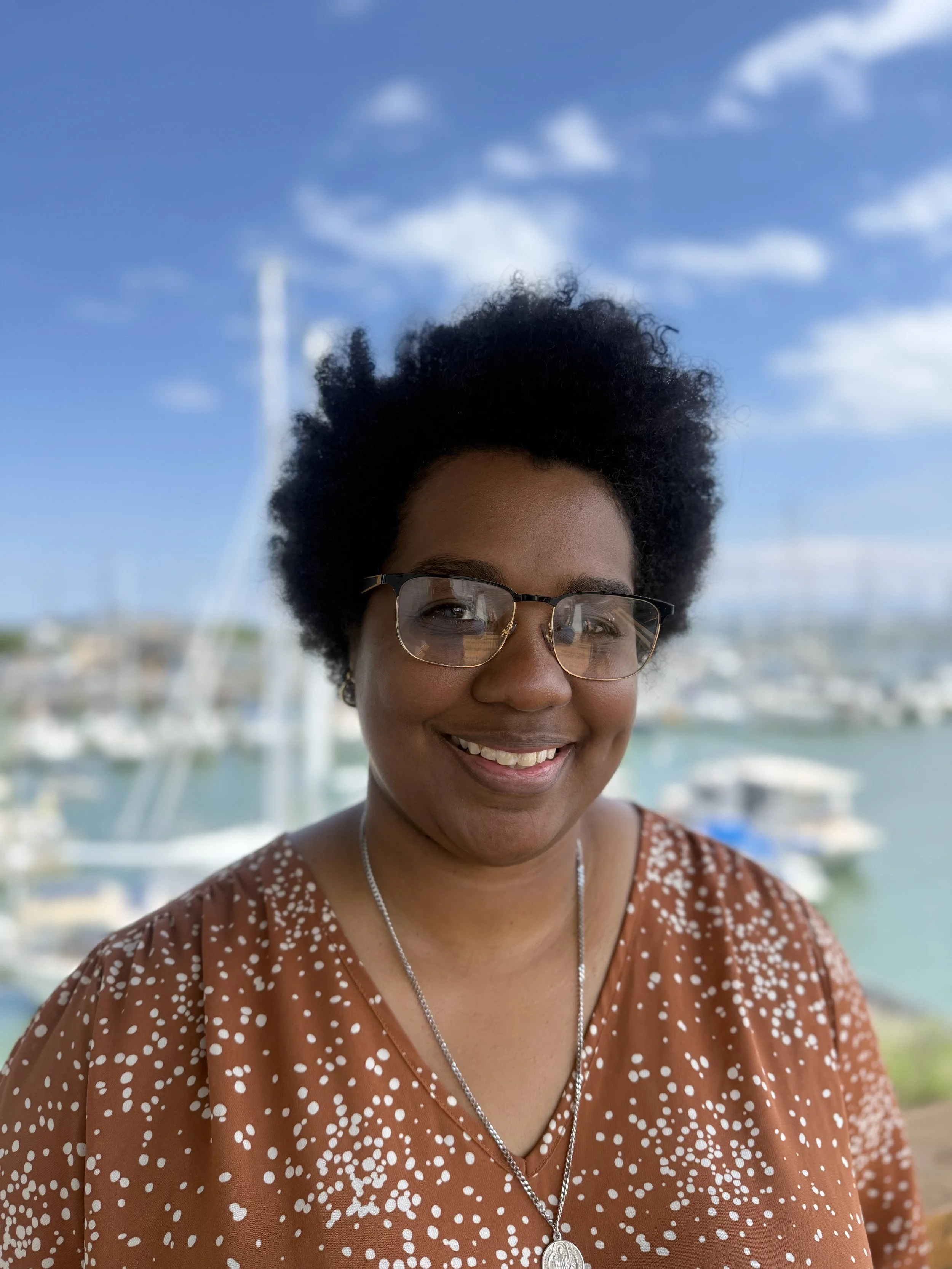 A smiling woman with short, curly black hair and glasses, wearing a brown shirt with white polka dots, standing outdoors near a marina with boats and a wind turbine in the background under a partly cloudy sky.