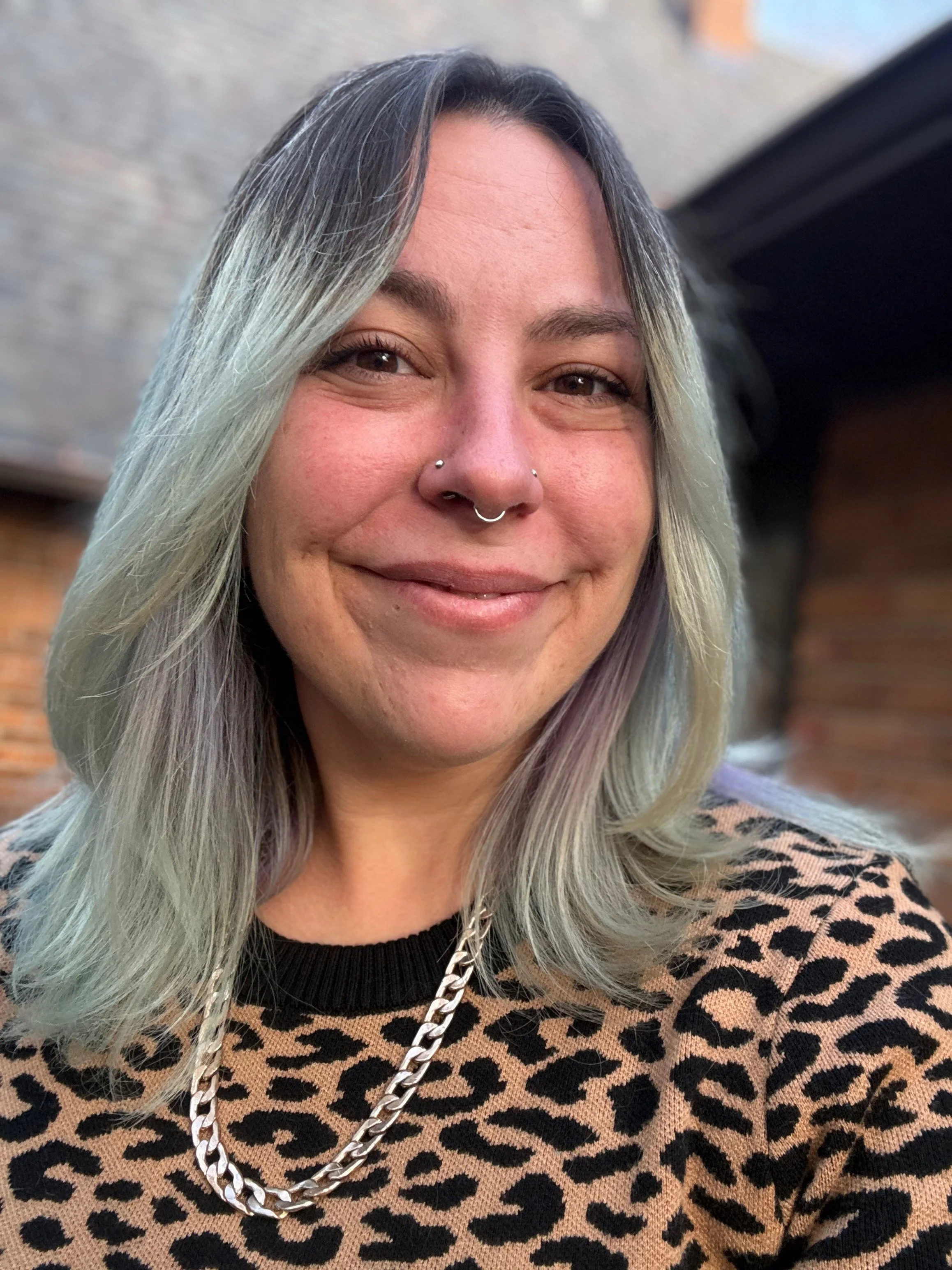 A woman with long, gray and blonde hair smiling at the camera, wearing a leopard pattern top and a chunky silver chain necklace, with a brick wall in the background.