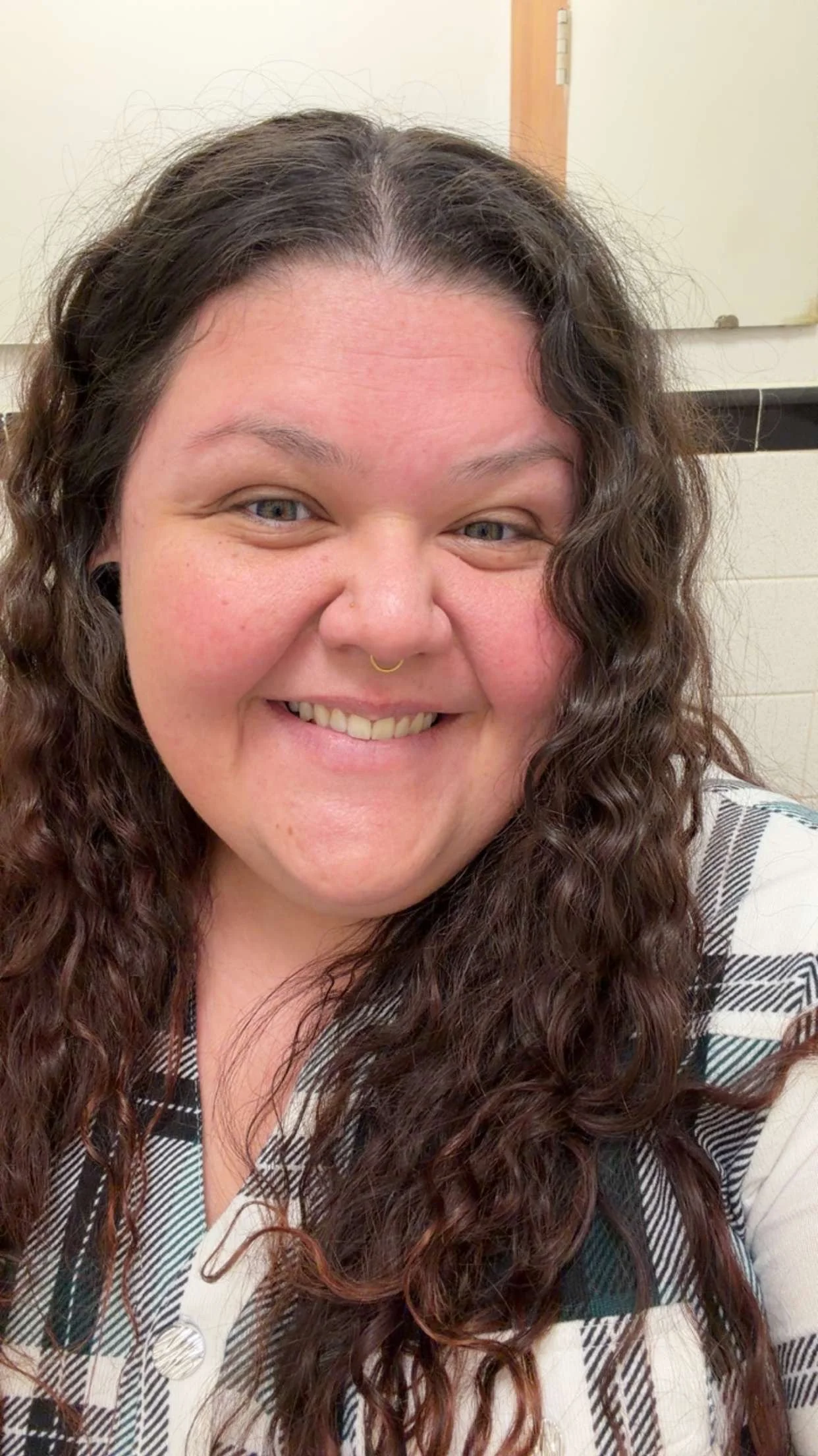 A smiling woman with curly brown hair, light skin, and gray eyes, wearing a nose ring and a black-and-white plaid shirt, in front of a beige wall with a wooden door.
