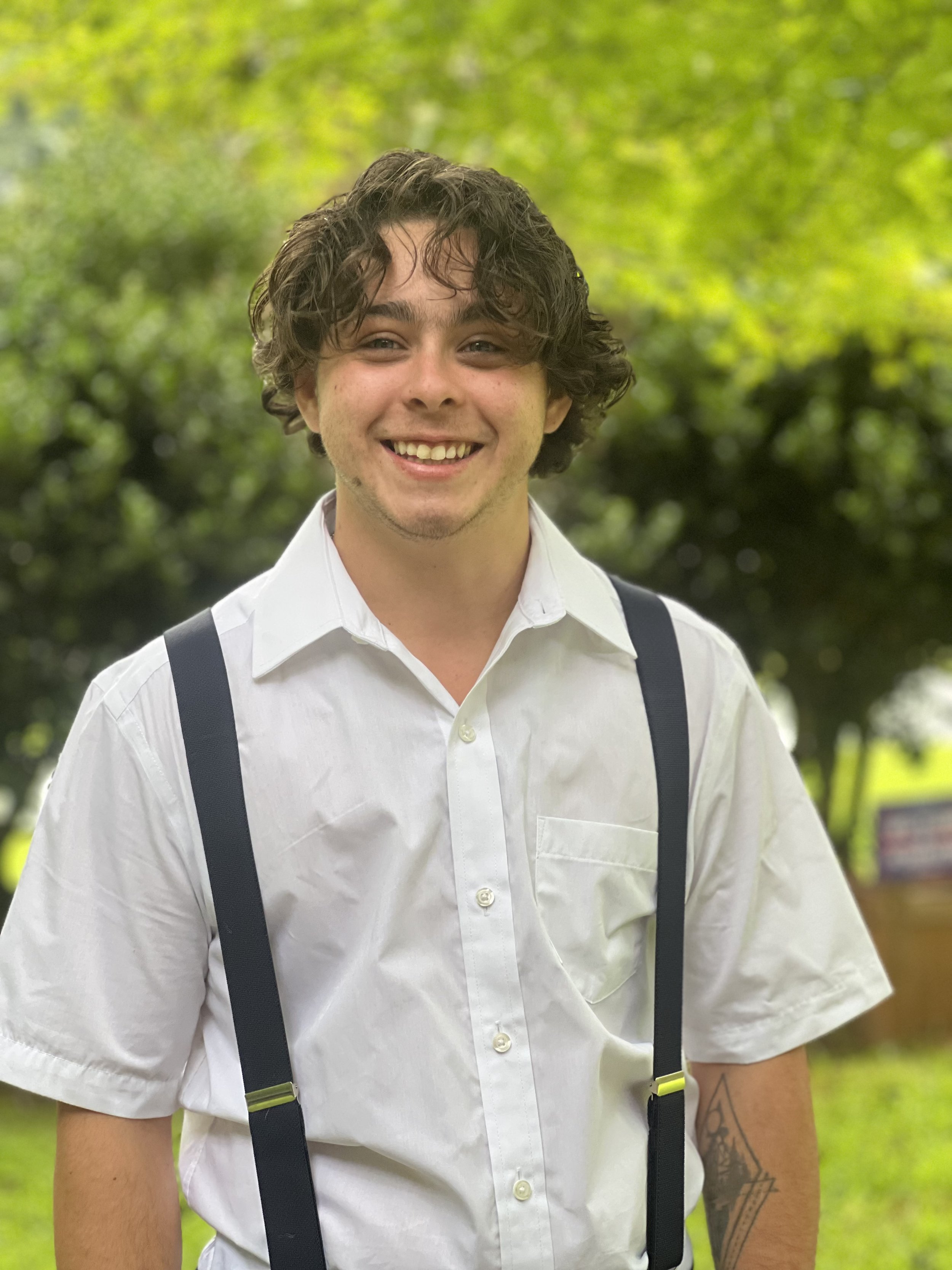 Young man with curly hair smiling, wearing a white shirt and suspenders, standing outdoors with green trees in the background.