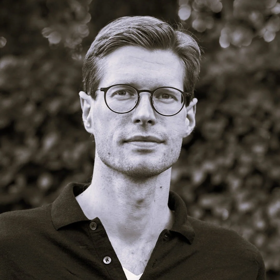 Black and white photo of a man with short hair and glasses, wearing a collared shirt, standing outdoors in front of a leafy background.
